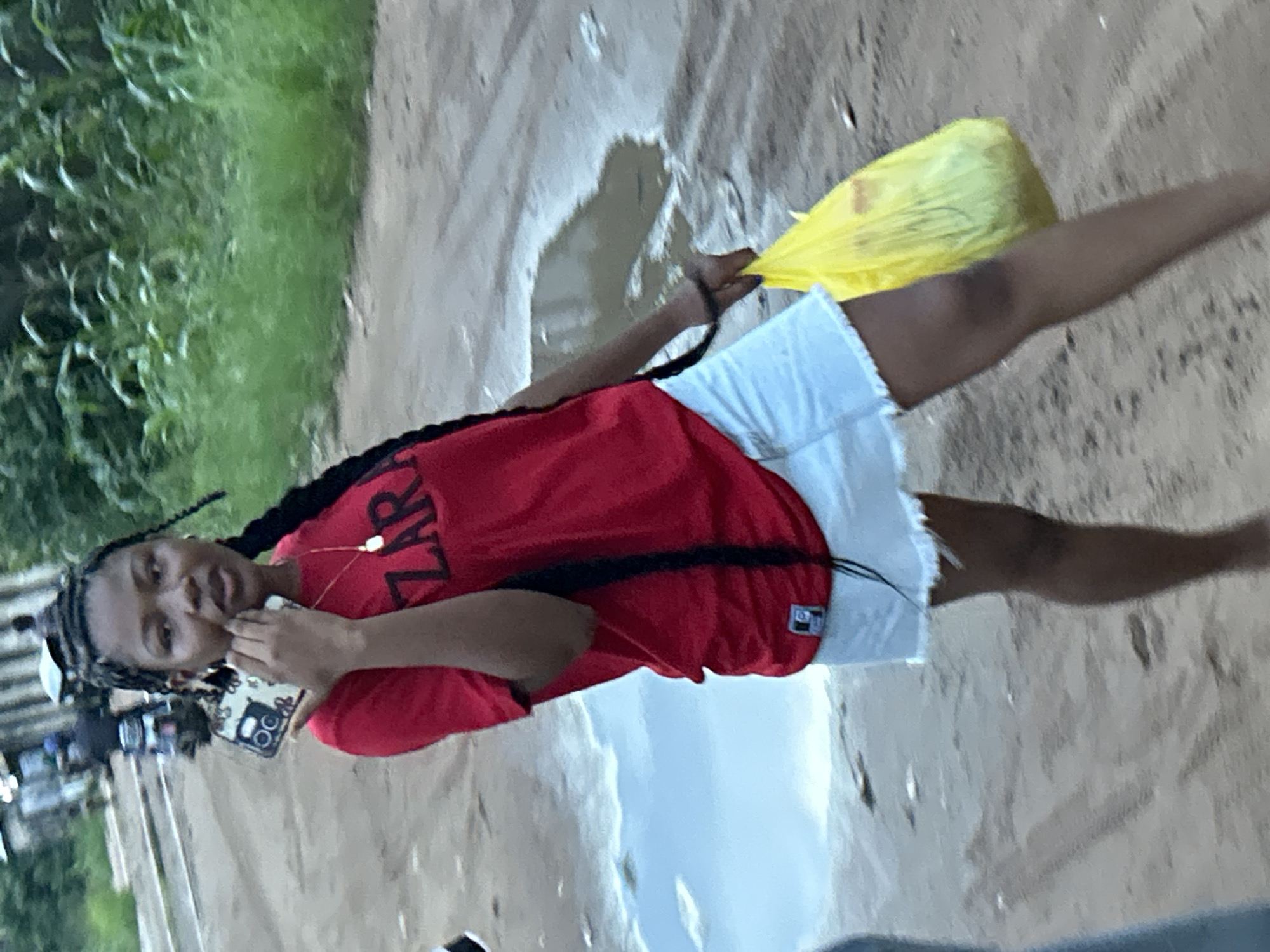 The image depicts a young woman standing outdoors in Ihiagwa, Nigeria. She has dark braided hair and is wearing a red short-sleeved t-shirt with "ZARA" printed in white capital letters on the front, paired with white shorts. White earphones with a visible cord are worn around her neck.
In her right hand, she holds a smartphone with a distinct black and white patterned case, brought close to her ear or face. Her left hand holds a bright yellow plastic bag, which appears to contain items, extended slightly to her side.
The setting features uneven, sandy, or muddy ground with visible puddles, suggesting recent rainfall. Lush green vegetation is present on the left side of the frame, indicating an outdoor, possibly rural or undeveloped area. The lighting appears soft, hinting at an overcast day, early morning, or late afternoon. The overall scene captures a casual moment in a damp, unpaved environment.