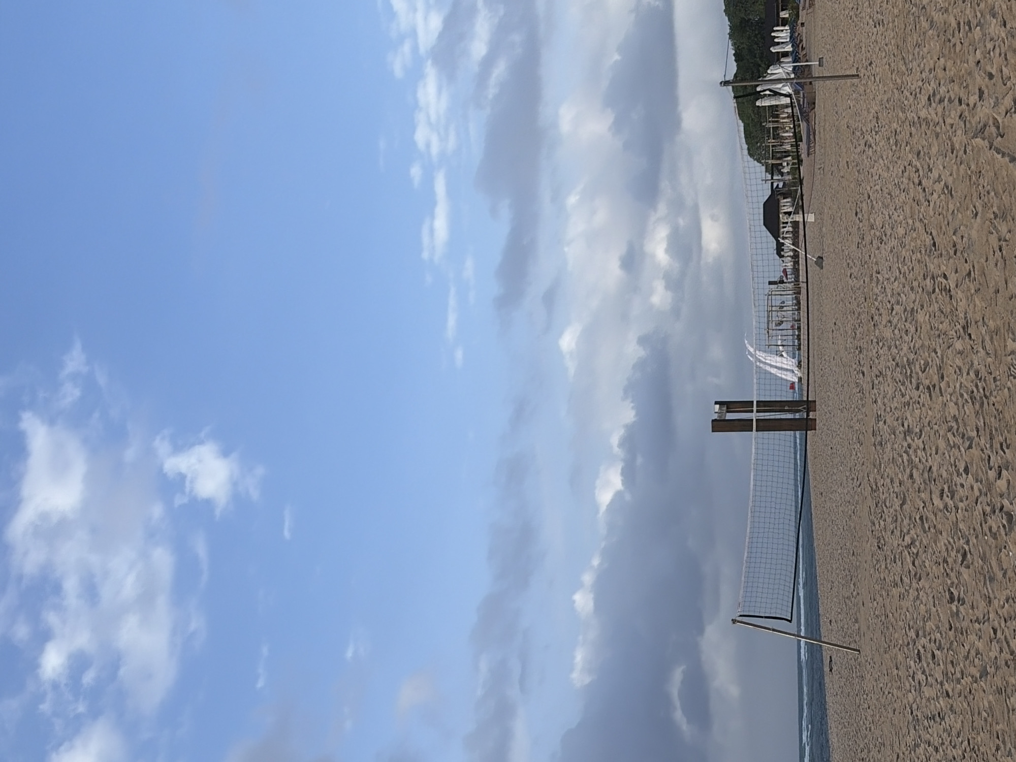 The image shows a beach volleyball net set up on a sandy beach. The ocean is visible in the background with waves. There are some buildings and umbrellas further down the beach. The sky is partly cloudy. This scene is consistent with beaches in the Kuta Selatan area of Bali, Indonesia, known for their tourist amenities and recreational activities.