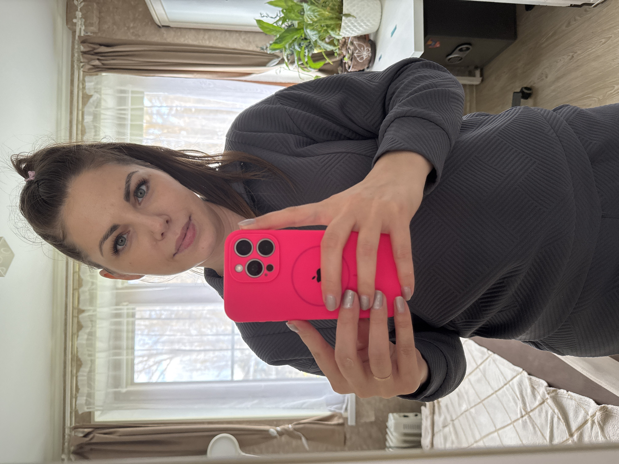 A mirror selfie captured indoors shows a young woman from the chest up, looking directly at the camera with a subtle, pleasant smile. The image is rotated 90 degrees clockwise. She has long, dark hair tied back, with a small pink hair clip visible, and light-colored eyes. She appears to be wearing makeup, including eyeliner or mascara.
She is dressed in a dark grey or charcoal long-sleeved top or dress, characterized by a textured geometric pattern. In her right hand, she holds a bright pink smartphone, which resembles an iPhone due to its triple camera array and a visible circular MagSafe-like design on the back, featuring an outline of the Apple logo. Her fingernails are painted a light grey or taupe color, and a thin gold-colored ring is visible on her left ring finger.
The background, reflected in the mirror, indicates an indoor setting, likely a bedroom or living area in Раздолье, Russia. A large window with white sheer curtains and brown blinds offers a view of bare trees outside, suggesting a cold season like late autumn or winter. The room features light-colored walls and a wooden floor. On a shelf or desk, a potted green plant with variegated leaves is visible. To the right, a bed with a white and grey patterned comforter is neatly made. Other reflected objects include a white rectangular device (possibly an air purifier or humidifier) and a dark box-like object on a shelf. The natural light from the window suggests it is daytime.