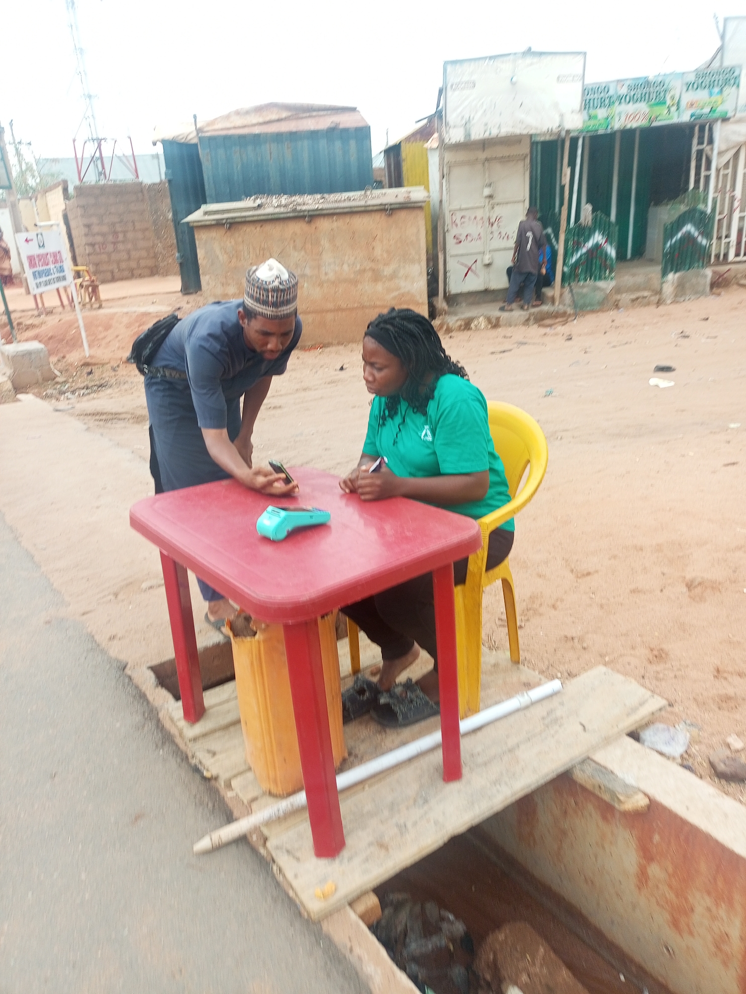 The image shows two individuals interacting at a red plastic table in an outdoor setting, likely in Gombe, Nigeria. One person, wearing a dark blue outfit and a patterned hat, stands and leans toward the table, holding a smartphone. A blue payment terminal is also on the table. The second person, seated in a yellow plastic chair, wears a green shirt and appears to be writing. The table is precariously positioned over an open storm drain covered partially by boards and a white pipe. Buildings and street signs are visible in the background, suggesting an urban or commercial area.