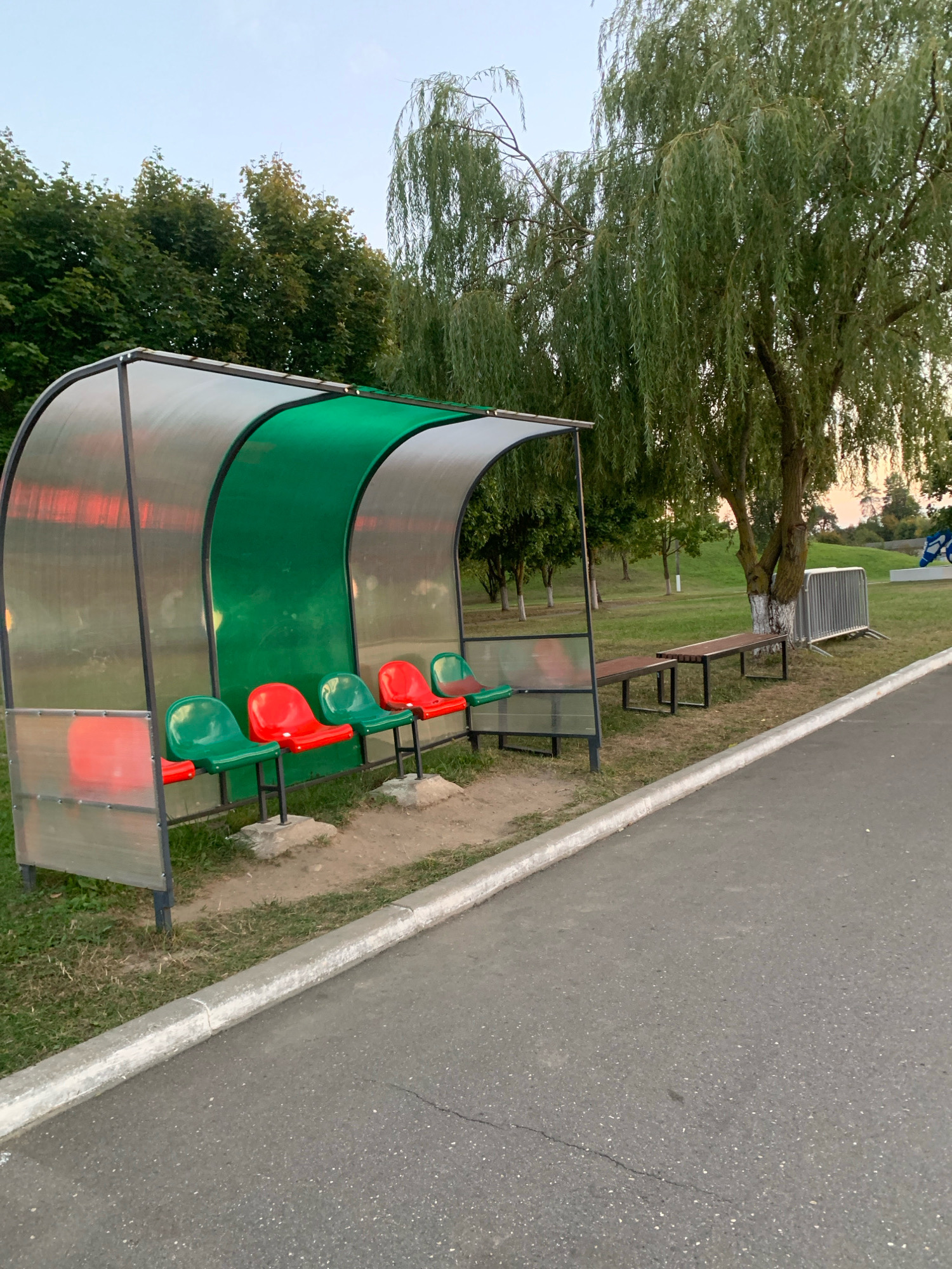 This is a medium-distance, eye-level, outdoor photograph taken in the city of Kapyĺ, Belarus. The image features a covered seating area, likely a dugout or shelter, situated next to a paved pathway. The shelter has a transparent, curved roof and a green back wall. Inside the shelter, there are several seats, alternating in red and green. In front of the shelter, there are two wooden benches. Behind the shelter, a large weeping willow tree stands prominently, its branches hanging low. The surrounding environment is grassy, with other trees visible in the background. The sky is overcast, suggesting it might be early morning or late afternoon. The scene appears to be a public recreational area, possibly a park or sports ground.