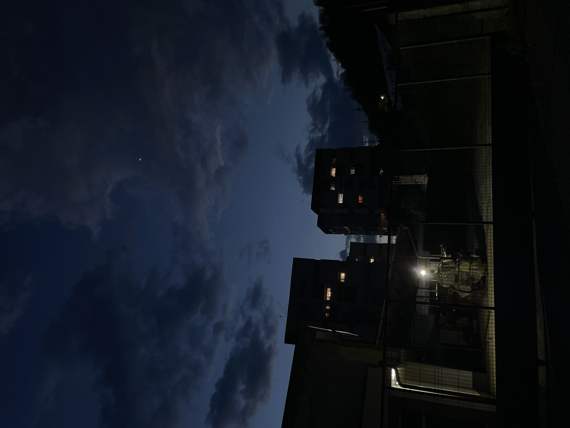 The image depicts a nighttime scene, likely in an urban area of Zlatograd, Bulgaria, based on the presence of apartment buildings. The sky is filled with dark, heavy clouds, suggesting an overcast condition. A small, faint light is visible high in the sky, potentially a star or a distant light source. Two apartment buildings dominate the middle ground, with a few windows illuminated. The foreground is dominated by a structure, potentially a fence or building element, with a bright light source shining on it, creating strong contrasts of light and shadow. The composition is oriented in a portrait format. The scene appears quiet and still, possibly in a residential neighborhood. The dark environment suggests limited ambient lighting.