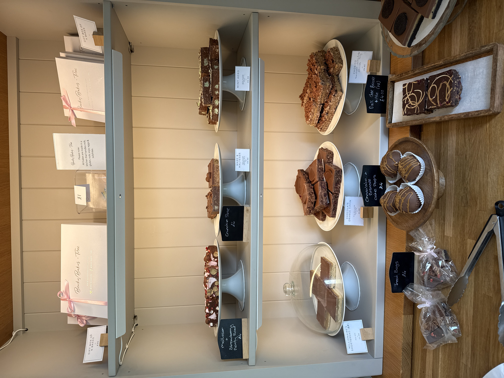 This image depicts a well-lit display case filled with various baked goods, likely located in a bakery or cafe in Unknown, Unknown. The case has multiple shelves, each displaying a different type of treat. From top to bottom, some items include "Mint Aero" bars, "Mars & Malteser" slices, "Crunchie Slice," and "Malteser + Strawberry Rocky Road." Additional items include "Chocolate Orange Tiffin," "Chocolate & Biscoff Cake Pops," "Dark Choc Biscoff Tiffin," and "Millionaire's Shortbread." Some items are on display stands, while others are individually wrapped in cellophane bags labeled "Treat Bags." Information cards with pricing accompany each item. The top shelf also features gift boxes from "Becky Bakes-Tree," along with a sign providing allergen information. A pair of tongs are visible on the right side of the frame. The background shows a light-colored wall with a simple design.
