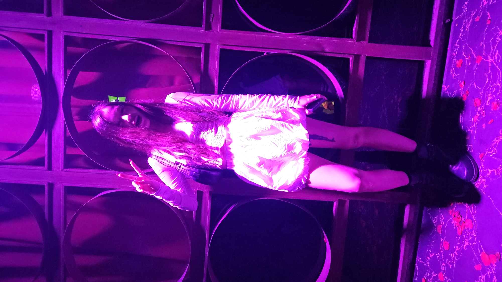 The image shows a woman in a white dress and black shoes, sitting on a wooden shelf with circular, dark objects, possibly mirrors or decorative elements, arranged behind her. The lighting is predominantly purple, creating a vibrant and somewhat surreal atmosphere. The background includes a wall with a marbled pattern, also illuminated in purple. The woman is making a peace sign with her fingers. A small, green rectangular object is visible above her head. Based on previous images and the mention of Nha Trang, Vietnam, this could be an interior scene, possibly a bar, club, or themed restaurant in Nha Trang. The arrangement of circular elements and the wooden structure suggests a specific aesthetic design characteristic of certain entertainment venues.