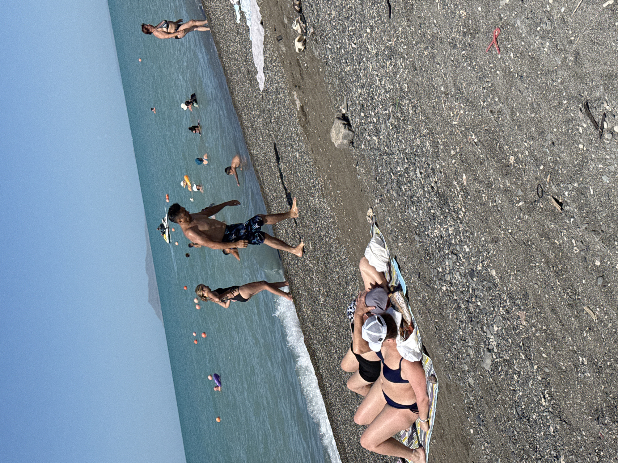 This image depicts a sunny day at a beach, possibly the shore of Lake Balkhash in Zharbulak, Kazakhstan. Several individuals are present in and around the water. In the foreground, two people are lying on beach towels on a pebbly shore, one wearing a dark blue bikini and the other with black shorts and a white and grey baseball cap. Further down the shore, a man in patterned swim trunks is walking away from the water.
In the shallow water, a woman in a black swimsuit is standing. A man in patterned swim trunks is walking in the water. Several other people are visible in the distance in the turquoise water, some swimming and others wading. A purple inflatable tube is in the water to the left. A jet ski is in the background on the water. The sky is clear blue, and a hazy landmass is visible on the horizon. The beach is covered in small, dark grey pebbles and sand. Scattered on the beach are some discarded items, including a pair of flip-flops and a red plastic object.