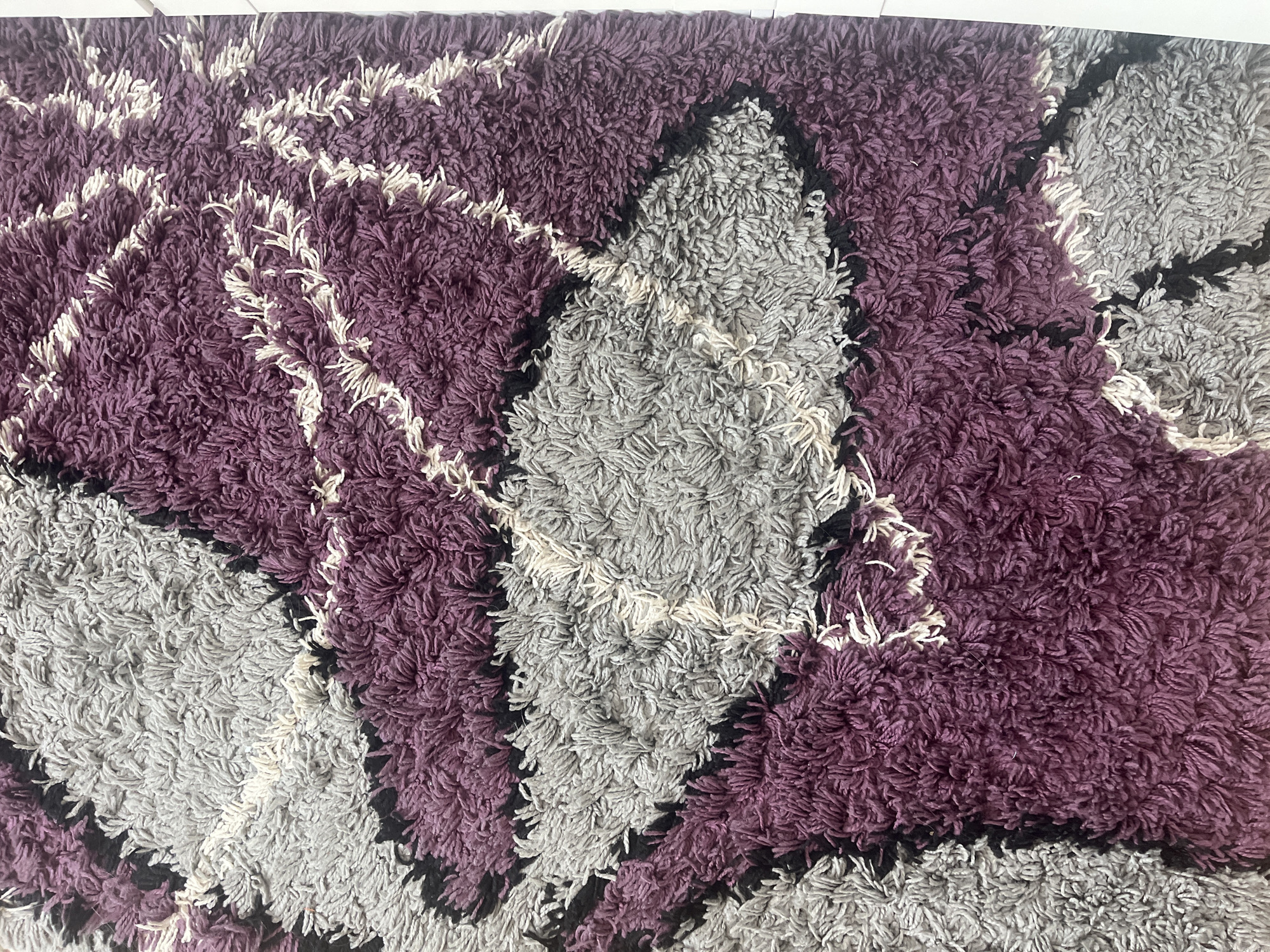 The image shows a close-up of a shag rug with a geometric pattern in purple, gray, black, and white. The texture is thick and shaggy. The pattern consists of alternating diamond-like shapes in purple and gray, separated by black outlines. White accents are scattered throughout the design. The image does not depict a location in Pokrovnik, Bulgaria, or any other identifiable location. There are no people present.