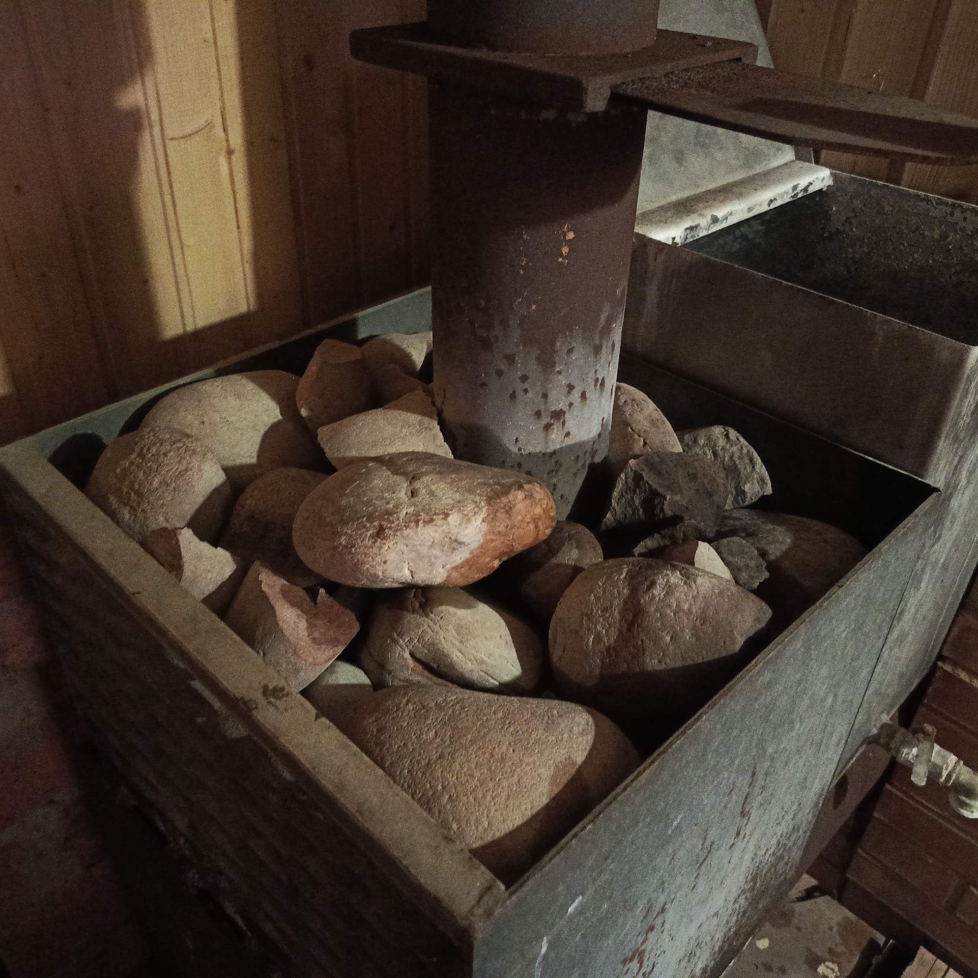 The image depicts a metal container filled with rocks, with a rusty metal pipe protruding from the center of the rocks. The container appears to be part of a heating system, possibly in a sauna or similar environment, as indicated by the wooden walls in the background. A water tap is visible on the side of the container. The location is Unknown, Unknown.