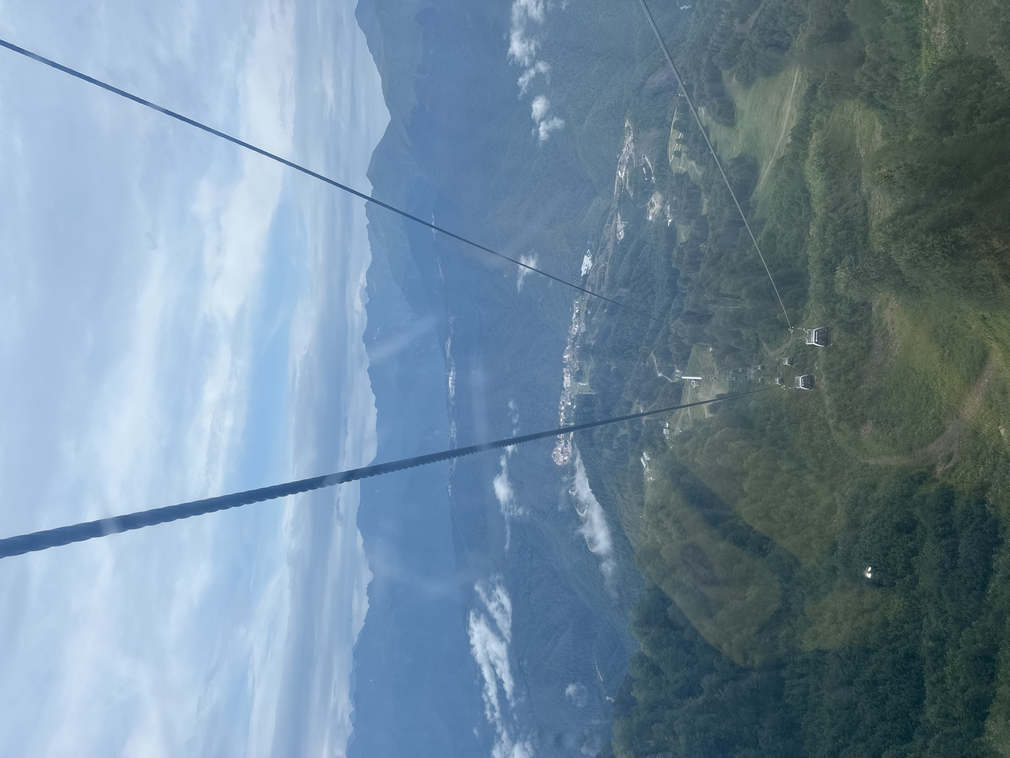 A wide-angle, eye-level shot captures a view from a cable car ascending a mountain range near Estosadok, Russia. The cable car's thick support cables dominate the foreground, angling towards the sky. Below, a vast expanse of lush green mountains stretches into the distance, punctuated by pockets of white clouds clinging to the slopes.
In the mid-ground, a small cluster of buildings, likely part of a resort or village, is nestled on a mountainside, with a winding road leading to it. Several cable car cabins are visible, traveling along the same cable system. The terrain is characterized by steep, tree-covered slopes, with some clearings and pathways visible. The sky is overcast, with patches of blue peeking through. The overall impression is one of a serene, natural landscape with human infrastructure integrated into it.