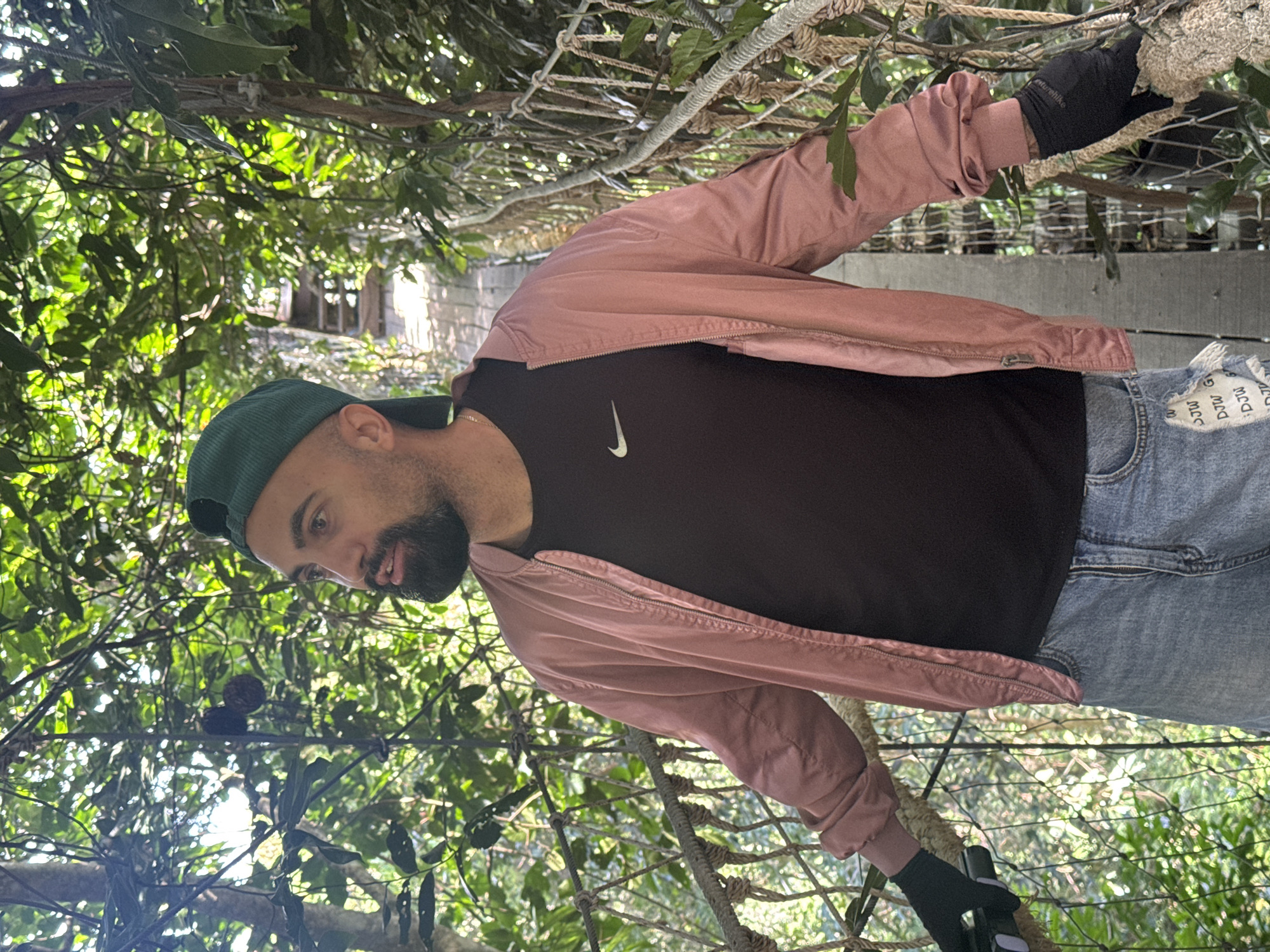 A man with a beard, wearing a green baseball cap, a pink bomber jacket over a black t-shirt with a Nike logo, and denim shorts, stands on a suspended walkway in a lush, green forest. He is wearing black gloves on both hands, with his left hand holding onto a rope railing. The walkway appears to be made of wood with rope railings and netting. The surrounding environment is dense with tropical foliage, and sunlight filters through the leaves. In the background, stairs are visible leading further along the walkway. The man is looking to his left.