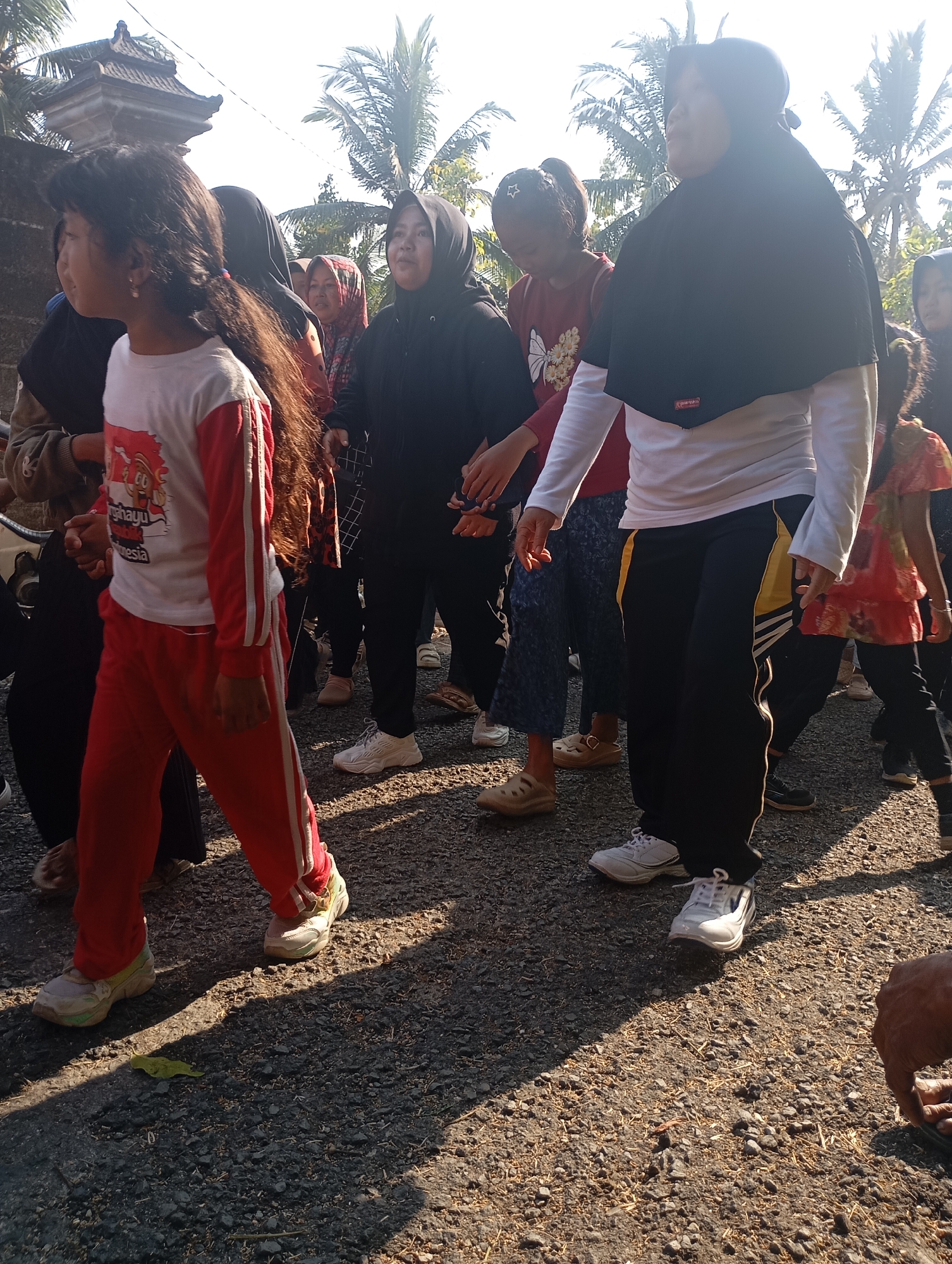 In Ketepung, Indonesia, a group of individuals, primarily women and girls, are gathered outdoors on a gravel surface. The lighting suggests it is daytime, with strong shadows indicating sunlight. Many of the individuals are wearing hijabs and modest attire, commonly seen in Indonesia. One young girl in the foreground is wearing a red and white long-sleeved shirt with a graphic and red track pants with white stripes. To her right, a woman in a black hijab and white long-sleeved shirt with black track pants featuring yellow stripes is visible. Other people are partially visible, some wearing colorful tops and dark bottoms. In the background, palm trees and a glimpse of a traditional Balinese-style roofed structure are present, suggesting a tropical or subtropical environment. The ground is covered in a mixture of gravel and dry vegetation. The overall scene appears to be a casual outdoor gathering, possibly a community event or a recreational activity.
