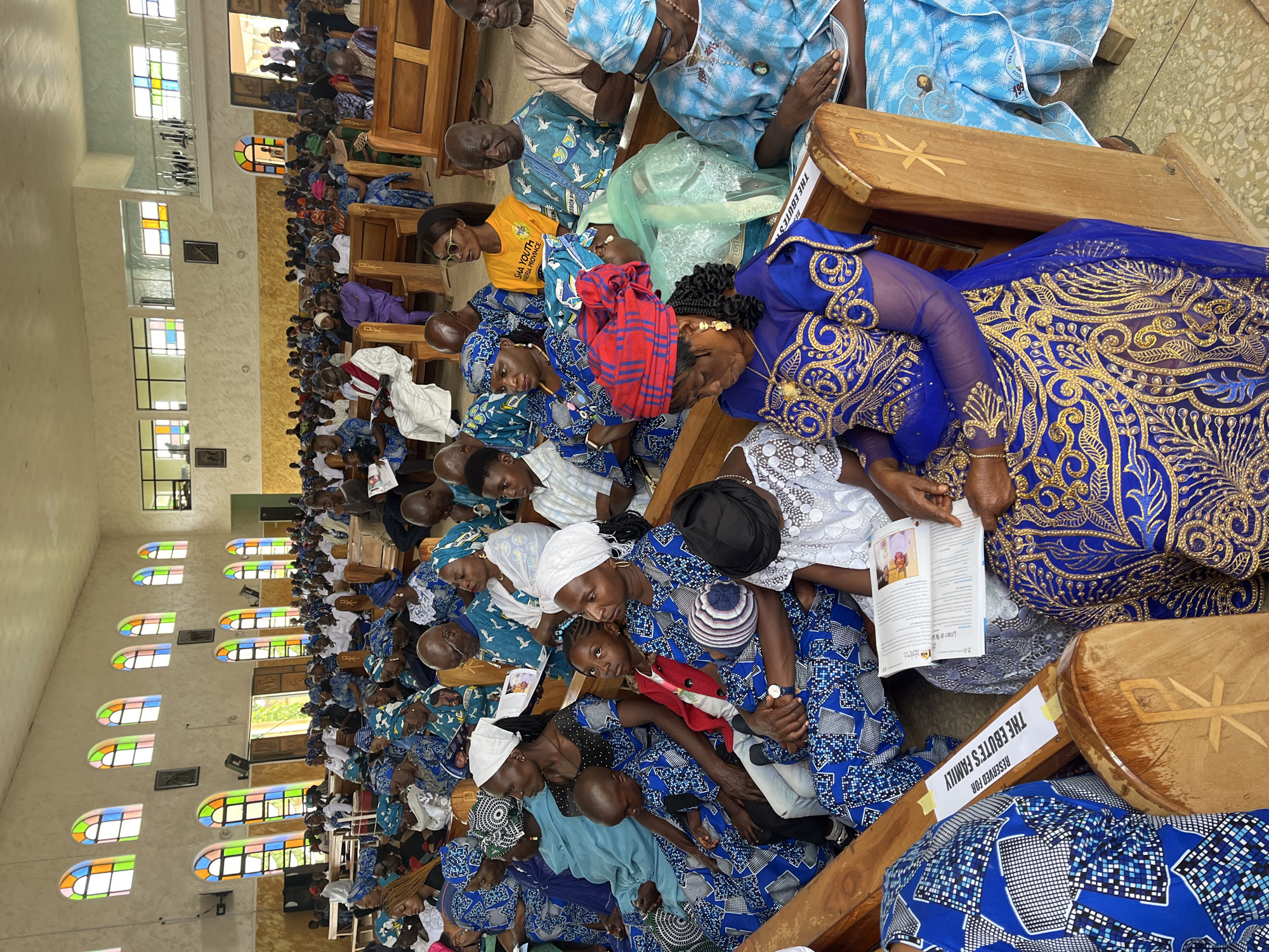 This is a medium shot, eye-level, full interior photograph of a church service in Vom, Nigeria. The church is filled with a congregation seated in wooden pews. The walls are a light beige color, and the ceiling is not visible. Above the congregation, on the far wall, are several arched windows with stained glass in various colors. The lighting is bright, likely from natural light sources.
The foreground of the image shows several people in the pews, predominantly women wearing matching blue and white traditional Nigerian attire with intricate patterns. Some are wearing headscarves in coordinating colors. A few individuals are holding prayer books or pamphlets. The central focus of the foreground is a group of women and children. One woman in a vibrant blue and gold dress with a matching headscarf is seated in the foreground, her head slightly bowed. To her left, a child in a blue and white patterned outfit is seated on her lap. Further to the left, another woman in a blue and white patterned dress is looking forward, holding a young child.
In the middle ground, the pews are filled with more congregants, men and women, also dressed in traditional attire. Some are looking towards the front of the church, while others are engaged in conversation or reading. A man in a light brown and white patterned outfit is seated in the middle ground, looking forward. A woman with a bright yellow shirt that reads "SHA YOUTH NIGERIA PROVINCE" is also visible.
The background shows more of the church interior, with additional rows of pews and congregants. The far wall of the church features a large arched doorway, through which sunlight streams in, revealing the exterior and some people standing. There are also framed pictures or icons on the walls. A wooden bench with a carved symbol that appears to be a Chi Rho is visible in the foreground. A piece of paper is taped to the pew that reads "RESERVED FOR THE EBUTE'S FAMILY". The floor is tiled with a speckled pattern.