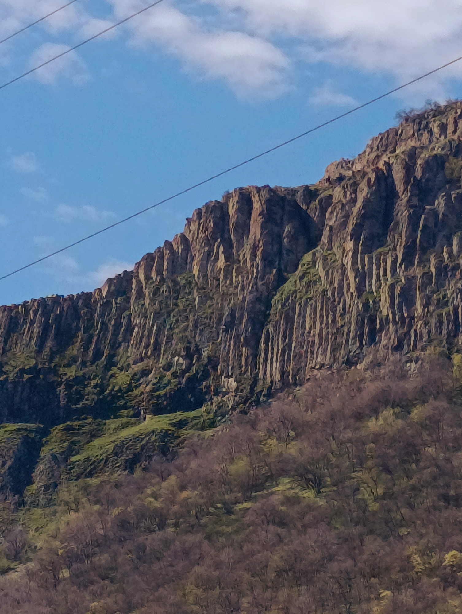 The image shows a steep, rocky cliff face, likely a geological formation of basalt columns, typical for volcanic regions. The cliff is partially covered with patches of green vegetation and bare trees at the base, suggesting early spring or late autumn. In the background, a blue sky with light clouds is visible. Two overhead power lines stretch across the upper portion of the frame. This is located in the city of Shiroko pole, Bulgaria.
