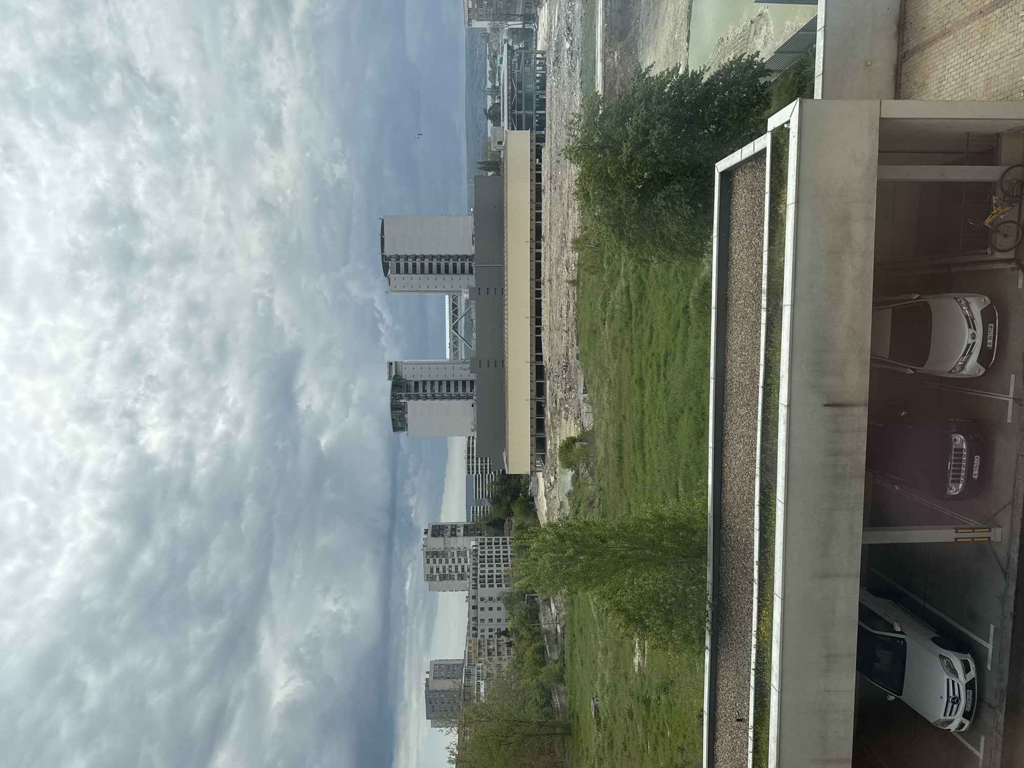The image presents a view from an elevated position overlooking an urban environment, possibly in Varna, Bulgaria. The sky is overcast with heavy cloud cover. The landscape features modern buildings of various heights and architectural styles. Construction or demolition work is visible near the water body in the distance. A green area with grass and trees is in the foreground. A building with a flat roof, a planted area, and a parking garage with vehicles parked inside is to the right. A bicycle is also parked inside the garage. The image appears to be taken during the daytime, with natural light.