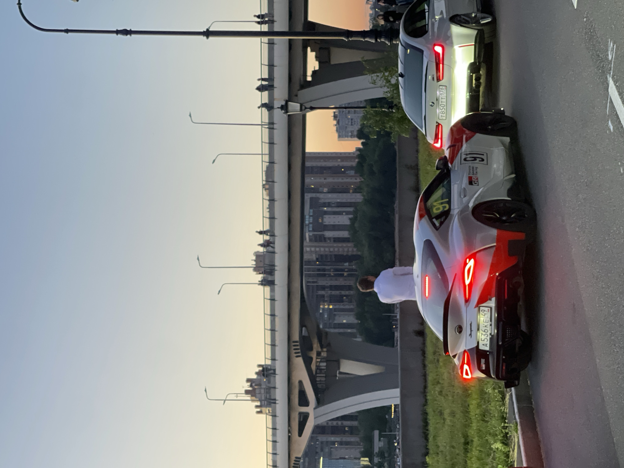 In the city of Unknown, Unknown, at dusk, a person in a white shirt stands with their back to the camera, looking towards a bridge. Two sports cars, a white and red Toyota Supra with the number 91 and a white BMW, are parked on a paved road in the foreground. The Supra's brake lights are illuminated. In the background, the illuminated bridge spans across a body of water, with pedestrians visible on its walkway. City buildings and trees are also visible under the bridge. The sky is a gradient of pale blue and orange, indicating sunset. Streetlights line the bridge and a separate lamppost stands to the right of the frame.