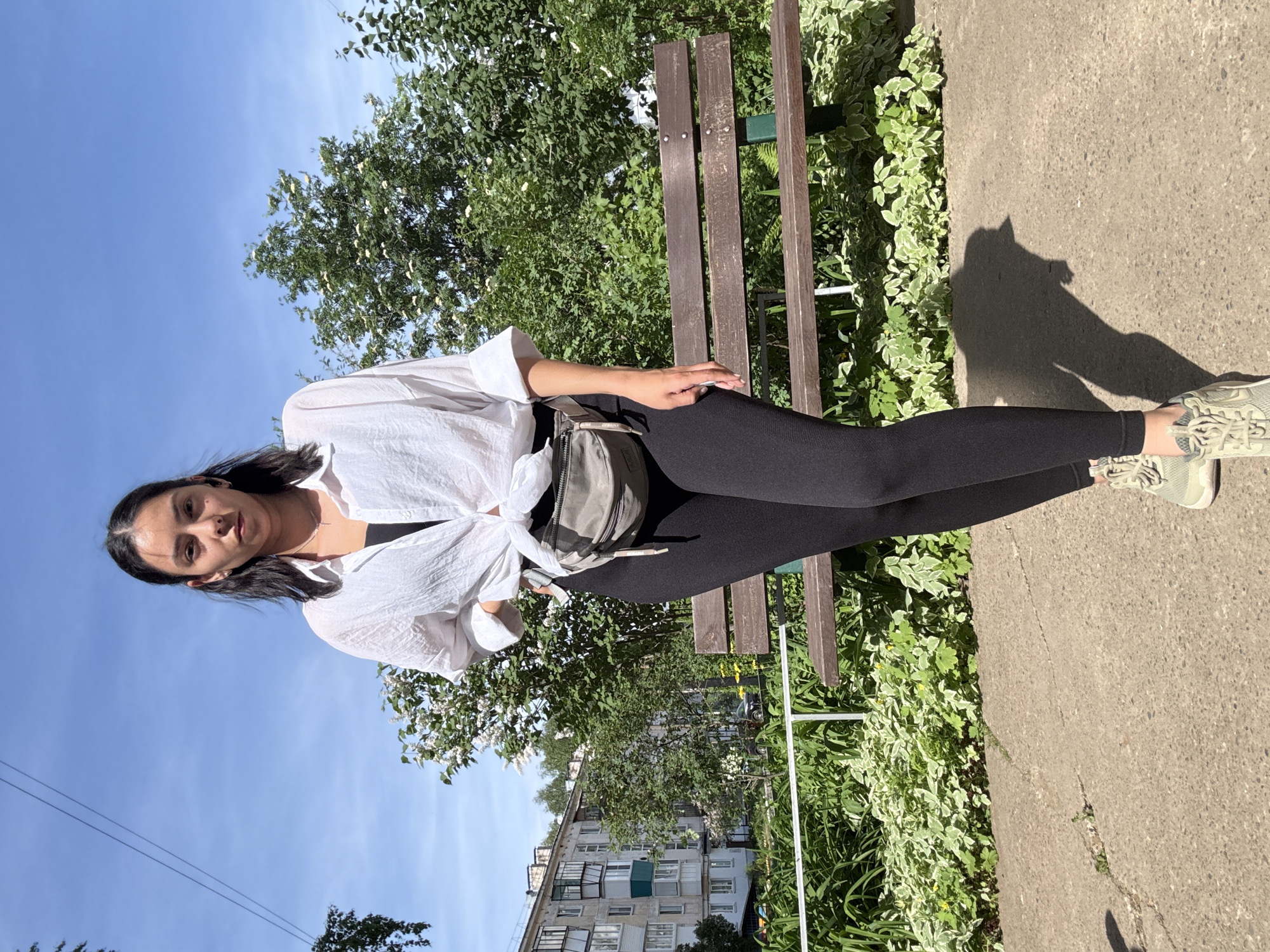 A young woman is sitting on a park bench in Chaikovsky, Russia. She is wearing a white shirt tied at the waist, black leggings, and light green sneakers. She is also wearing a gray fanny pack. The bench is made of dark wood and metal. There are trees and bushes behind the bench. A sidewalk is visible on the right. A multi-story building is visible in the background. The sky is blue.