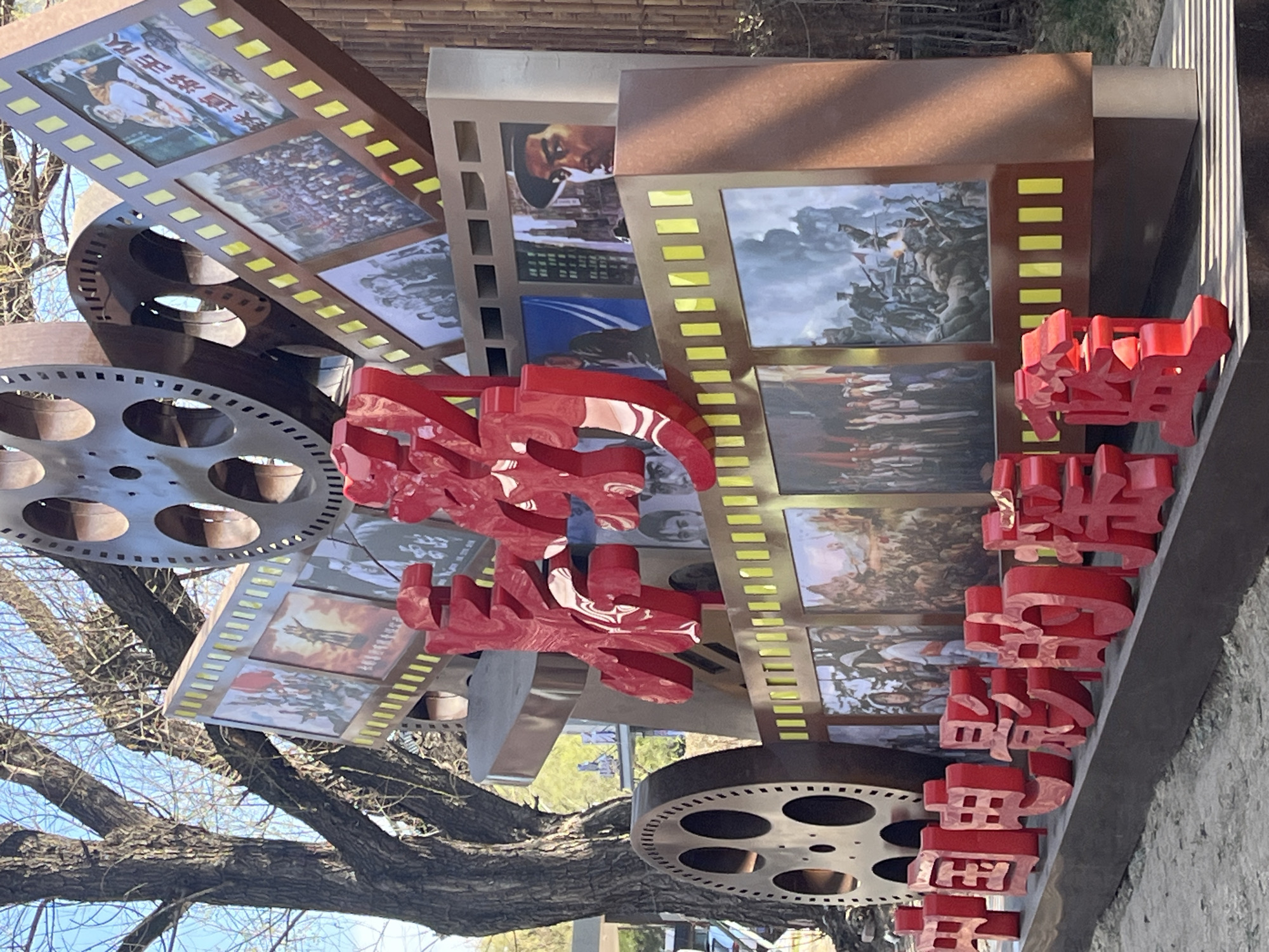 The image shows a street art installation in Changchun City, China. The installation features film reels and strips of celluloid that appear to be constructed from metal, composite materials, or plastic. The film strips are mounted with stills from historical films. Overlaying the film elements are large, three-dimensional red Chinese characters, possibly forming a phrase or a name related to Chinese cinema. The installation is located outdoors, as suggested by the presence of a bare tree on the left side. The ground is a cement pathway. The scene is in daylight.