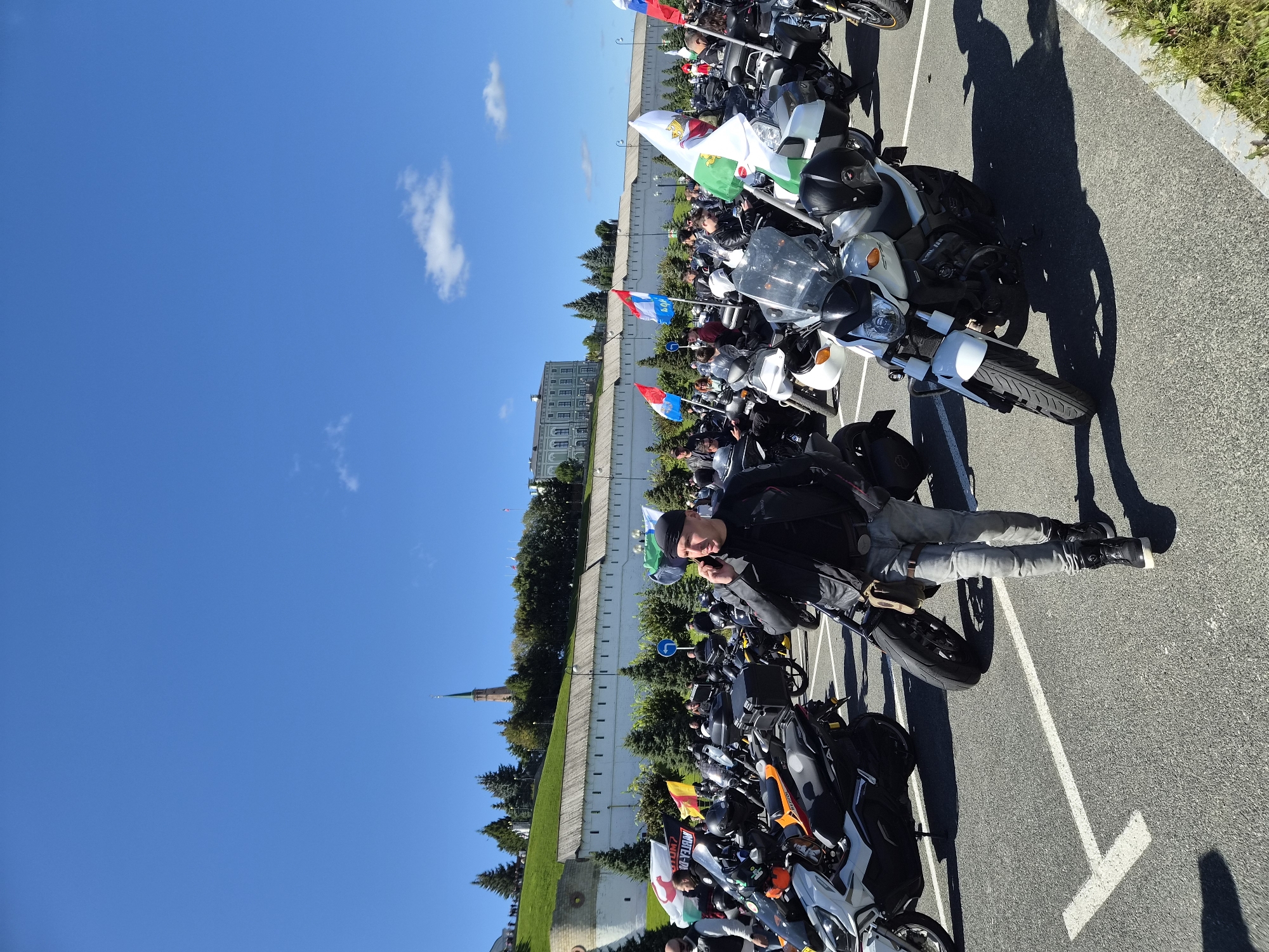An outdoor scene depicts a gathering of motorcycles and people in a parking lot. A man in a black leather jacket, black bandana, and grey jeans is walking and talking on his phone. He is positioned in the foreground, with several motorcycles parked around him. In the background, a white wall stretches horizontally, topped with trees. Behind the wall, a light green building with a spire is visible, likely part of the Kazan Kremlin. The sky is a clear blue with a few white clouds. The image appears to have been taken in Kazan, Russia, given the architectural style and potential context of a motorcycle rally.