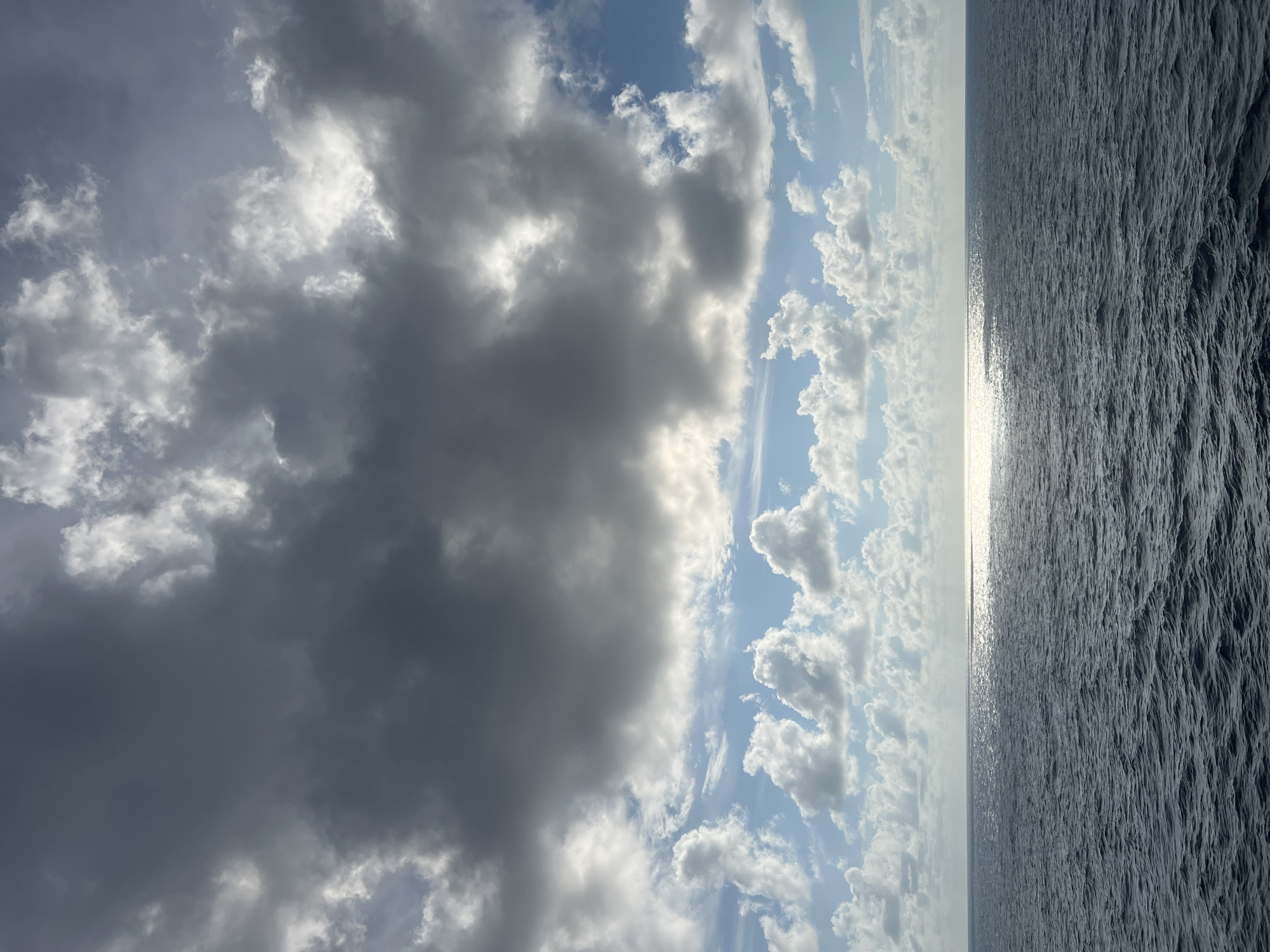 The image shows a vast expanse of dark blue ocean meeting a sky filled with a mixture of dark and light clouds. The sun is partially obscured by the clouds, but its rays are breaking through, creating a shimmering reflection on the water's surface. The clouds in the upper portion of the image are heavy and gray, suggesting an impending storm or the aftermath of one. Below these, lighter, fluffier clouds are scattered across a bright blue sky. The sea appears choppy, with visible waves. The scene is devoid of any human presence or man-made structures, evoking a sense of isolation and the raw power of nature. The location is described as Unknown, Unknown, indicating that specific geographical details are not available.