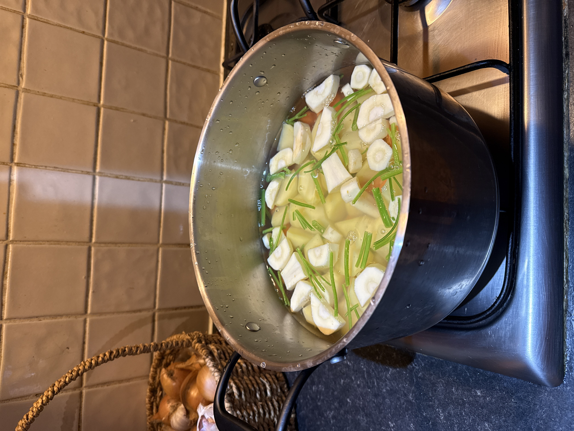 A medium-angle shot captures a pot of food on a stovetop, situated in what appears to be a domestic kitchen. The pot, made of stainless steel and black material, contains water, diced potatoes, sliced parsnips, and chopped chives. The pot is positioned on a modern, black stovetop with stainless steel trim. Adjacent to the stovetop, a wicker basket overflows with onions and garlic. The background reveals a tiled kitchen wall with light brown, square tiles. The kitchen setting suggests a home environment. The city of Great Yarmouth, United Kingdom, cannot be conclusively determined from the image.