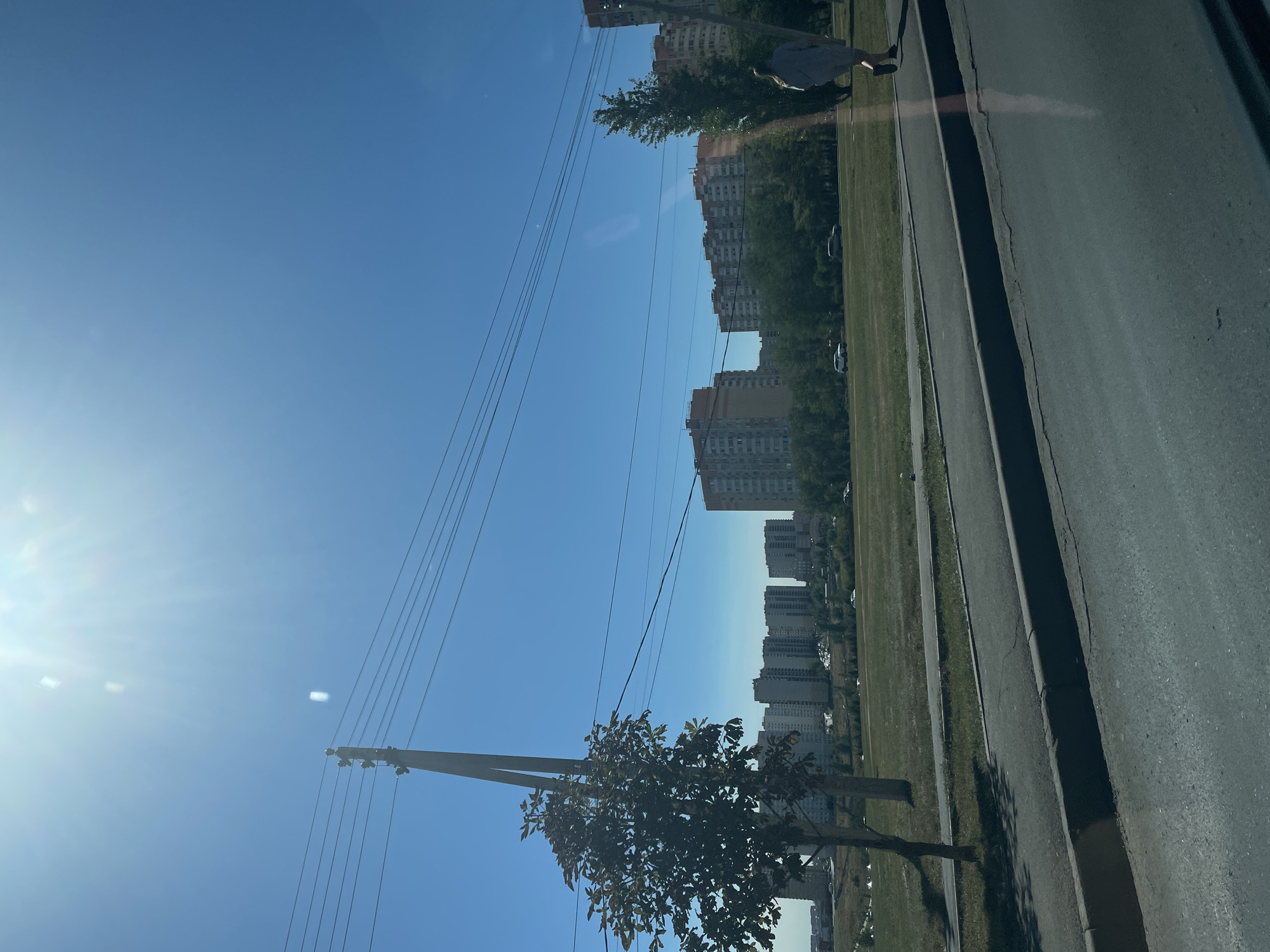 In the city of Rostov-on-Don, Russia, on a clear day with bright sunlight, a street scene unfolds. A paved road occupies the foreground, bordered by a curb and a sidewalk. Beyond the sidewalk, a grassy area stretches out, dotted with trees. In the middle ground, a series of multi-story apartment buildings forms the urban backdrop, their facades displaying a range of colors and architectural styles. Utility poles with numerous wires are visible, suggesting the presence of infrastructure. A solitary person is walking on the sidewalk, their back to the camera, dressed in a light-colored garment. The overall impression is of a typical urban environment under a bright, cloudless sky.