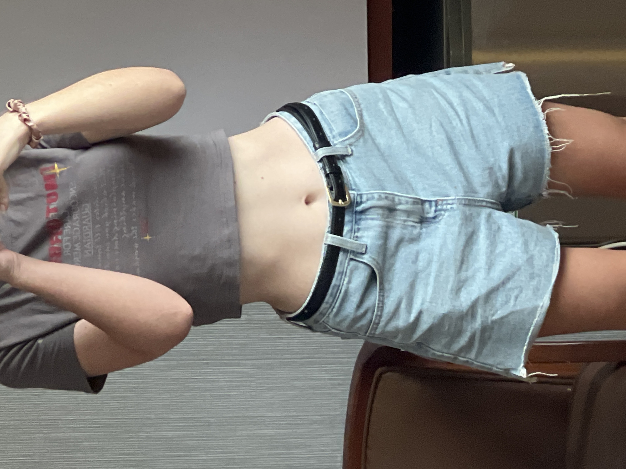 A person is wearing a gray t-shirt and light blue denim shorts with a black belt. The person is indoors with a gray wall in the background. A wooden chair is partially visible. The exact location within Bac Nha Trang Ward, Vietnam cannot be determined from the image.