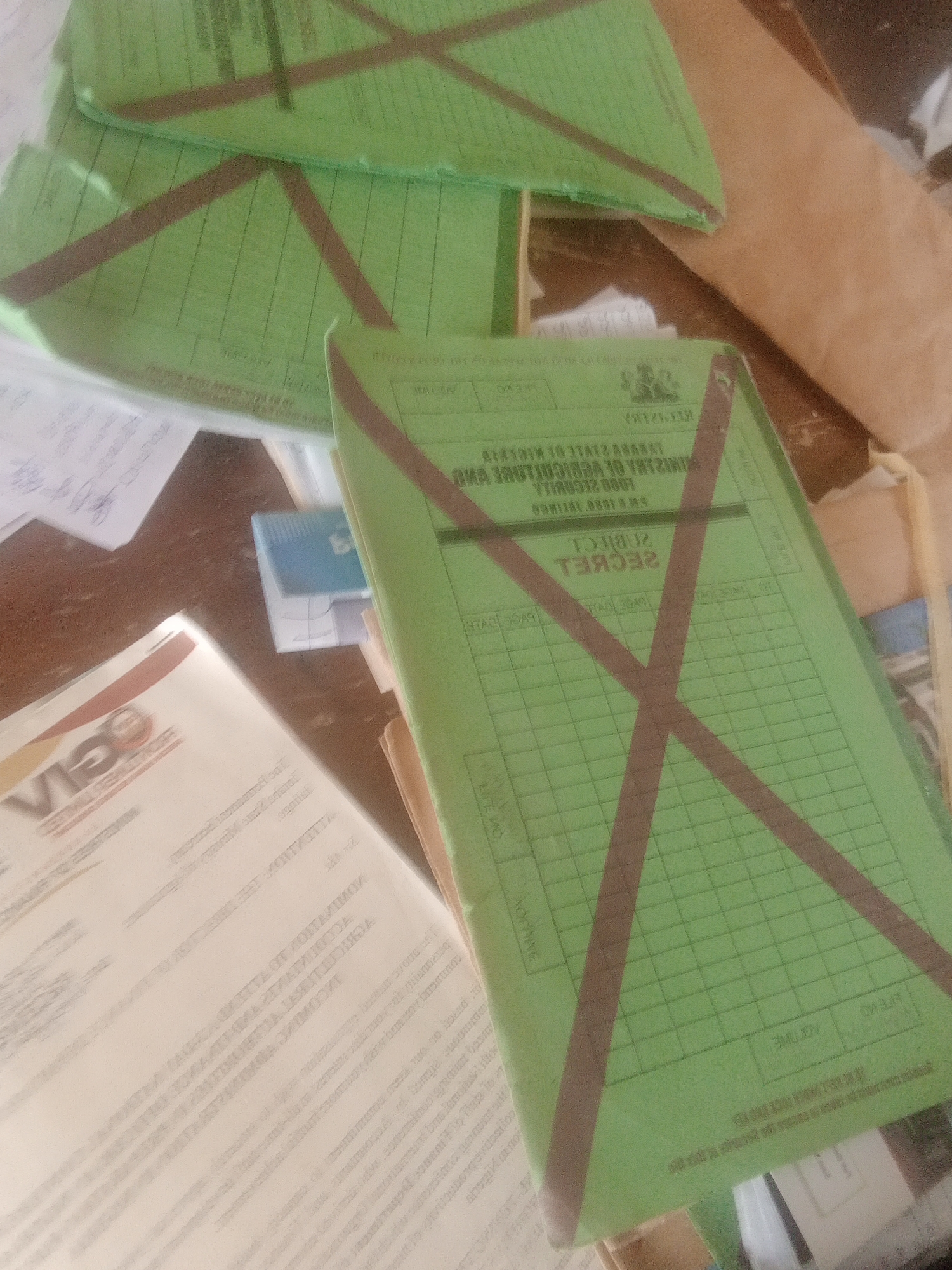 The image shows a stack of green folders, some with brown tape forming an X across them. The folders appear to be official documents, with text printed on them that is partially visible. One folder on the right has the word "SECRET" printed on it in large letters. Another folder on the left has a logo and text that is not fully legible. The folders are on a wooden surface, and there are other papers and objects scattered around. The scene appears to be an office or storage area. The context of Jalingo, Nigeria is not evident from the image itself.