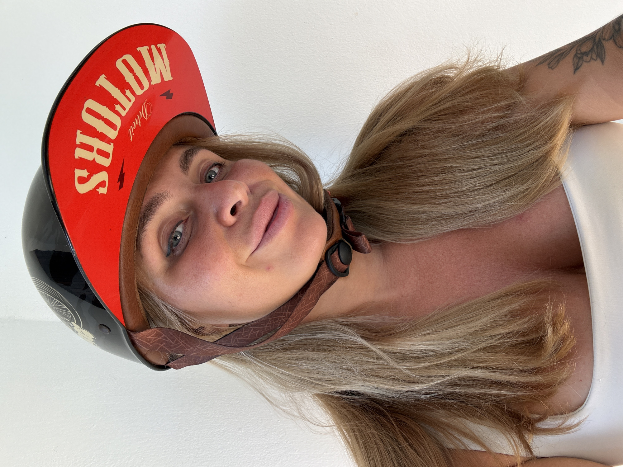 A blonde woman with fair skin and freckles is looking at the camera and smiling. She is wearing a black and red motorcycle helmet that says "SHOLOW MOTORS DETROIT" on the brim. Her hair is long and blonde, and her chest is visible, with some cleavage showing. She is wearing a white top. The background is a white wall. The image appears to be taken indoors, possibly in a home or studio. There is no indication of the location being in the city of Ko Samui District, Thailand.