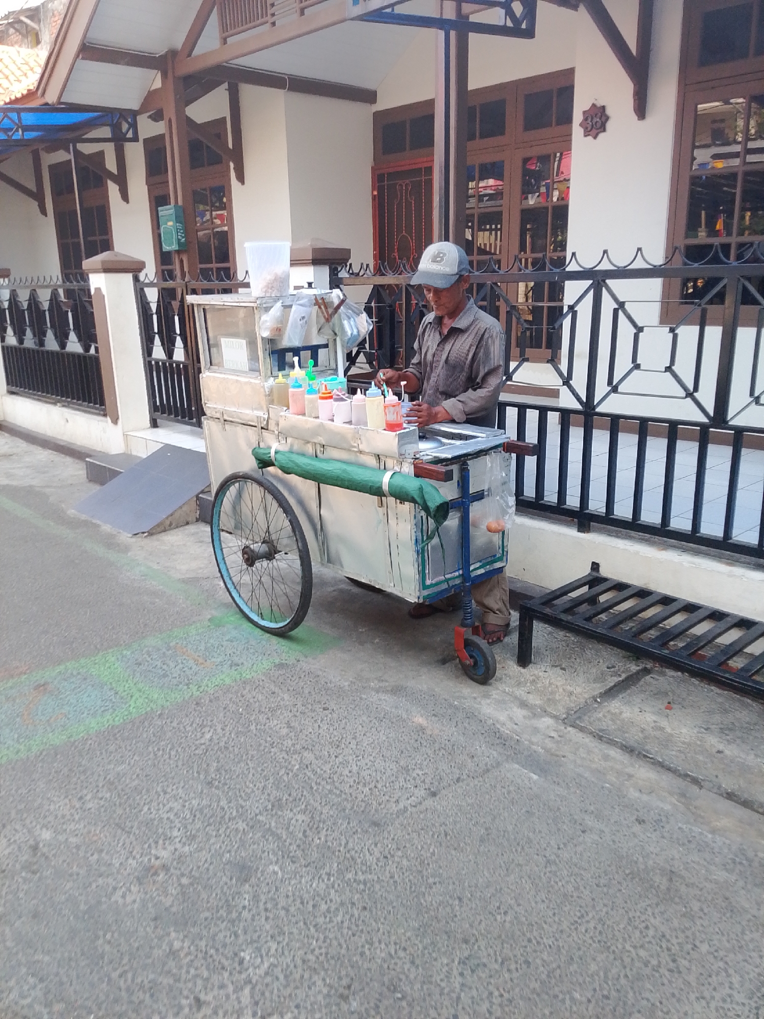 A man stands behind a food cart on a street in East Jakarta, Indonesia. The man, wearing a grey striped shirt, a grey baseball cap, and brown pants, is preparing food. The food cart is silver and has a large wheel on the left side and two smaller wheels on the right. It is equipped with bottles of condiments, a clear container, and plastic bags of food. The cart is positioned in front of a house with a white facade, brown window frames, and a wooden porch. A black metal fence encloses the property. The street is paved with asphalt and has some green markings on it. A metal bench is visible to the right of the cart. The lighting suggests it is daytime.
