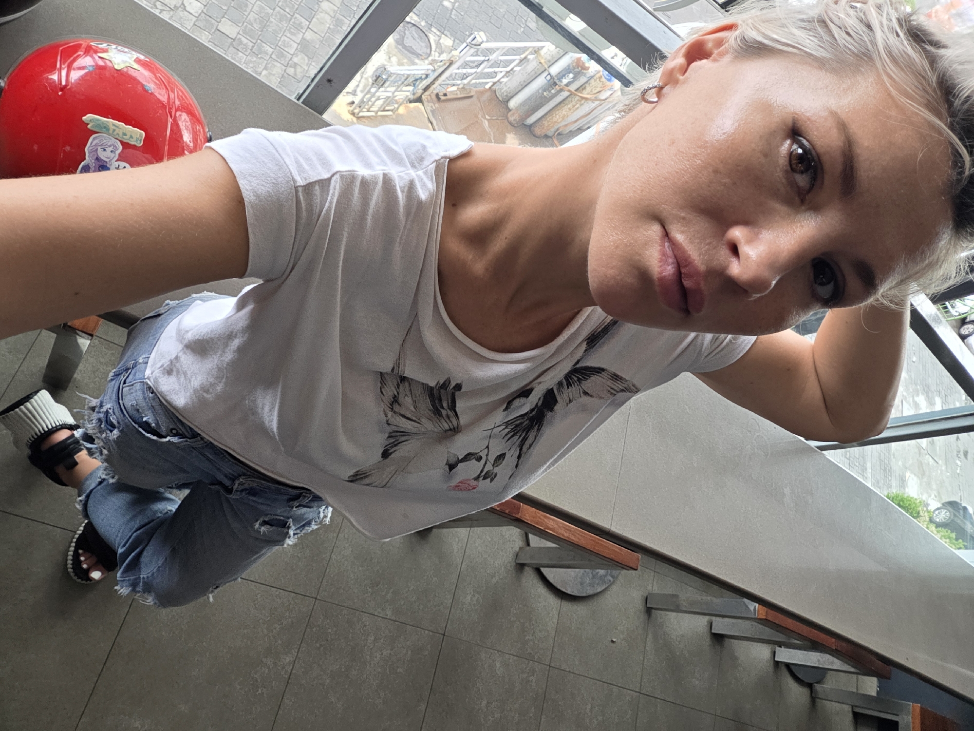 A high-angle selfie photograph shows a fair-skinned woman with short blonde hair, wearing a white t-shirt with a graphic bird design and ripped blue jeans, sitting at a table. The woman's face is close to the camera, capturing her facial features. She has a serious expression. Her right hand is behind her head. She is wearing an earring. She is wearing open-toe sandals. Beside her, there is a red helmet with stickers, including one featuring a character from "Frozen". The table is gray. The location seems to be an indoor space with a view to outside, possibly Kuta, Indonesia. The setting gives a glimpse of urban surroundings with structures visible through the windows. The gray tile flooring suggests a public or commercial space.