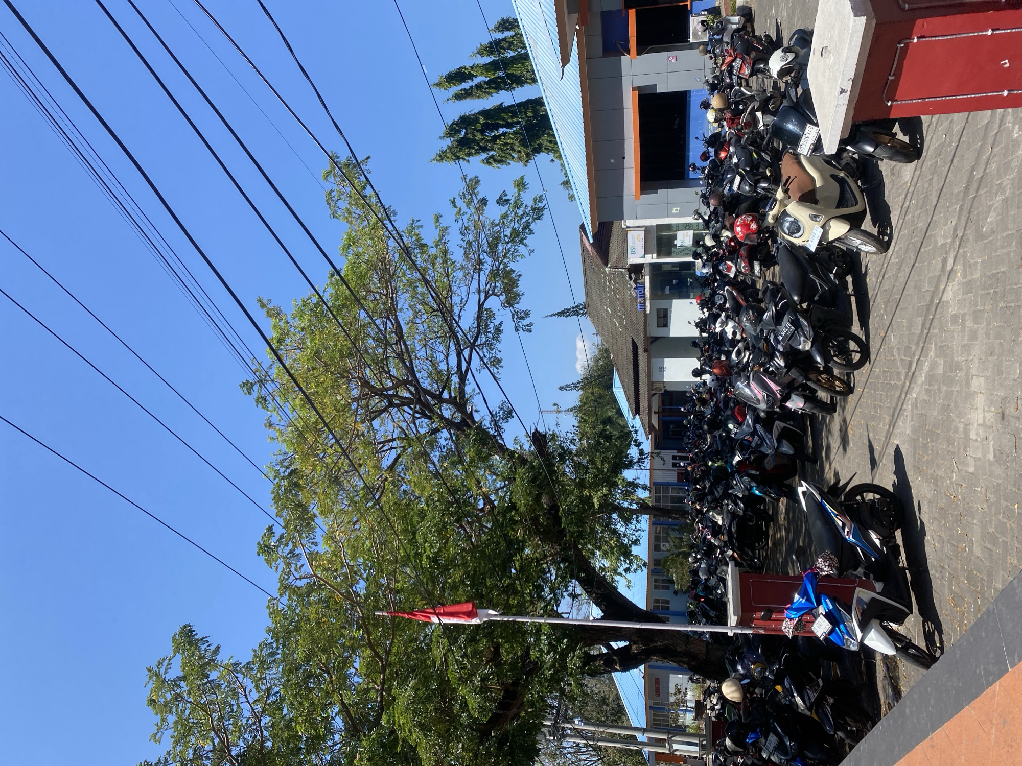 The image depicts an outdoor scene in Bantaeng, Indonesia, likely a public space such as a parking lot outside a government building or bank. A large number of motorcycles are parked closely together on a paved surface. In the foreground, a bright blue and white scooter stands out. A tall tree with lush green foliage occupies the left side of the frame, with an Indonesian flag flying from a flagpole near its trunk. Behind the parked motorcycles, a row of low-rise buildings with light blue roofs is visible. The sky above is clear and bright blue, with several power lines crisscrossing the upper portion of the image. The overall impression is one of a sunny day in a busy urban environment.