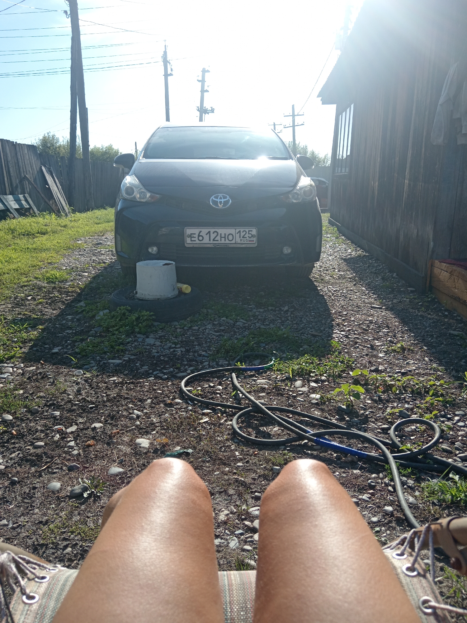 The image depicts an outdoor scene in Novopokrovka, Russia, on a sunny day. In the foreground, the legs of a person are visible, sitting in what appears to be a hammock or chair. Their legs are tanned and appear to be covered in fine hair. The person is wearing patterned shorts. Behind the person, a dark-colored Toyota car is parked on a gravel and dirt path. The car's license plate, E612HO 125, is clearly visible. A black hose is coiled on the ground near the car, and a concrete cylinder rests on a tire next to the vehicle. To the right of the car, a rustic wooden structure, likely a shed or building, is present. A wooden fence runs along the left side of the scene, separating the yard from an overgrown area with trees and grass. Overhead, power lines are strung between wooden poles. The sunlight casts strong shadows across the ground.