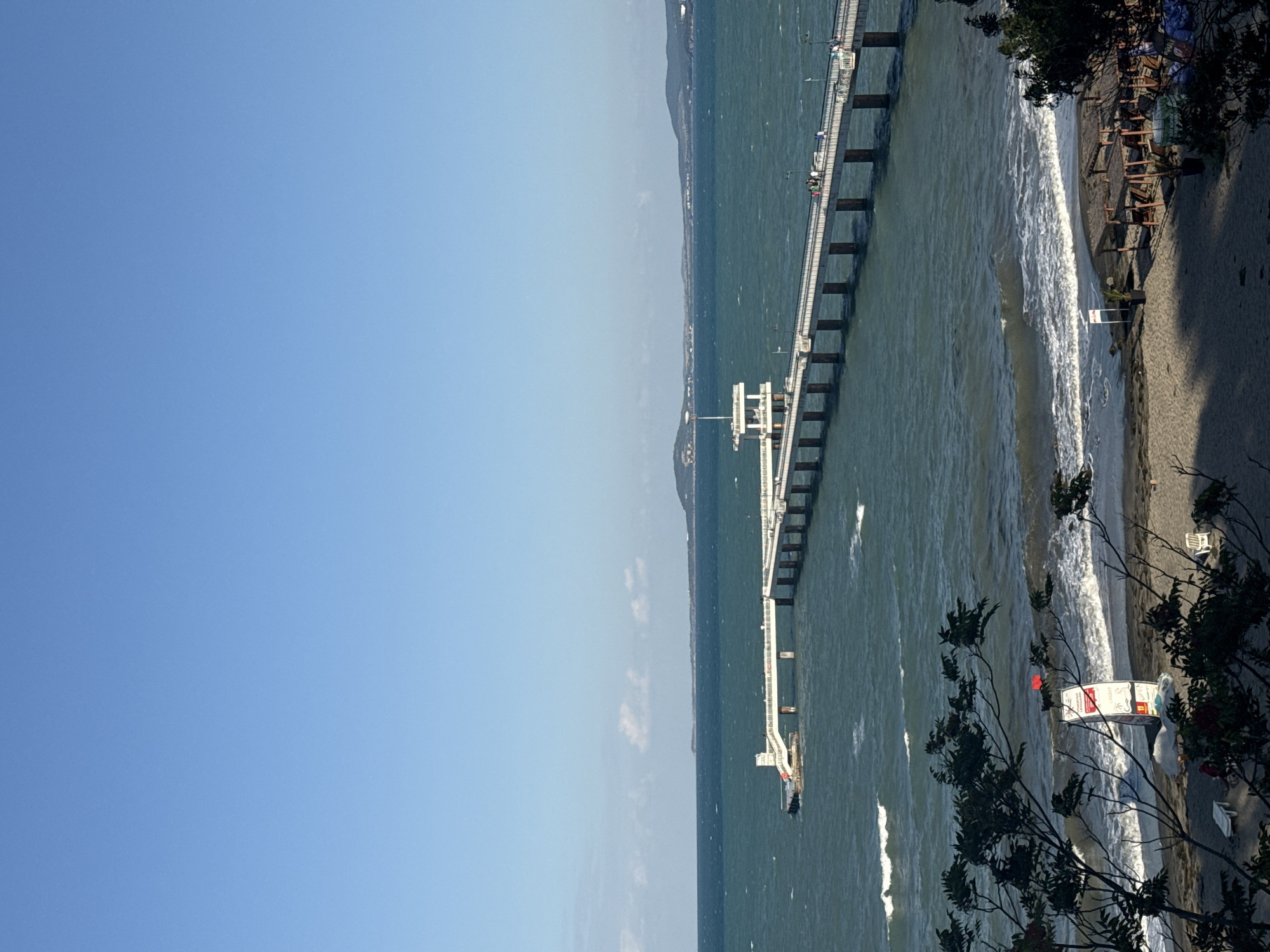 The image shows a pier extending into the sea, with a beach and trees in the foreground. The pier is white with railings and lamps. There are a few people on the pier. The sea is blue and choppy, with waves breaking on the shore. The sky is clear and blue. The beach has sand and some beach chairs and umbrellas. The trees are leafy and green. The scene is likely the Burgas Sea Garden in Burgas, Bulgaria, known for its long pier.