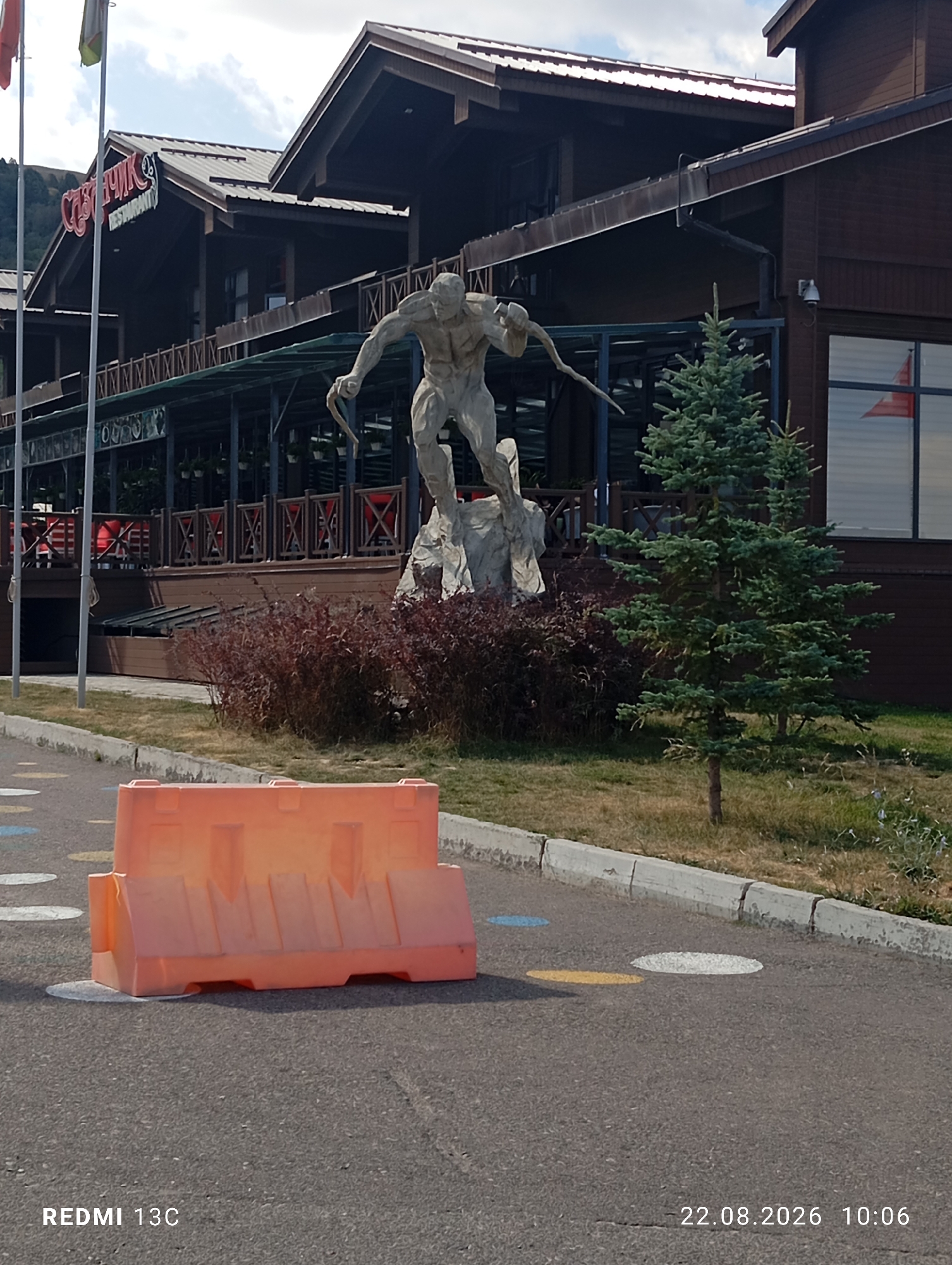 This is a full-shot, eye-level outdoor photograph taken at the Amirsay ski resort in Uzbekistan. The scene depicts a large, multi-story wooden building with a dark brown exterior and a dark-colored roof. A prominent statue of a muscular, sculpted human figure is positioned in front of the building, holding a spear-like object. A small evergreen tree stands to the right of the statue. In the foreground, an orange plastic barrier is placed on a paved surface, which is marked with several circular painted dots in yellow, white, and blue. The sky is visible in the upper portion of the image, with scattered clouds. The overall environment appears to be a well-maintained outdoor area, possibly a public space within the resort. The bottom right corner of the image displays the timestamp "22.08.2026 10:06" and the text "REDMI 13C". There are no people visible in the image.