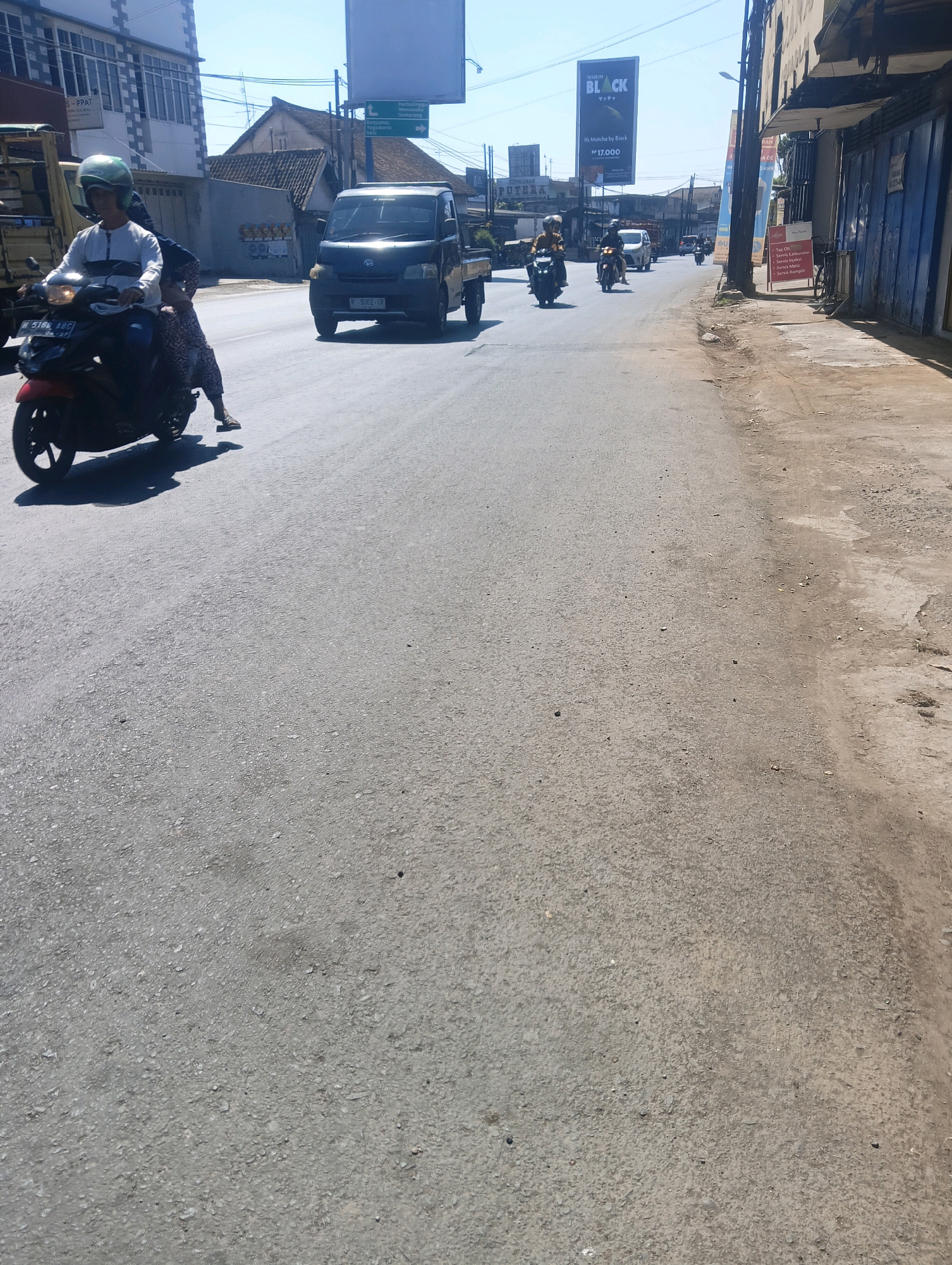 The image depicts a street scene in Sokaraja Kidul, Indonesia, under a clear blue sky. In the foreground, an asphalt road occupies the majority of the frame. On the left side of the road, a red motorcycle is in motion, carrying two individuals. The rider, wearing a green helmet and a white and black long-sleeved shirt, is in focus. Behind the rider, a person wearing a patterned fabric is seated. The motorcycle casts a shadow on the road.
Further down the road, a dark-colored pickup truck is visible, with its license plate reading "R 9062 CR". Several other motorcycles are also seen traveling in the same direction. On the right side of the road, commercial buildings with blue facades and signage are present. A black billboard with the word "BLACK" prominently displayed, along with Japanese text and currency symbols, stands on the right side of the road. Other advertisements and poles are also visible along the street. The environment suggests a commercial or residential area with active traffic.