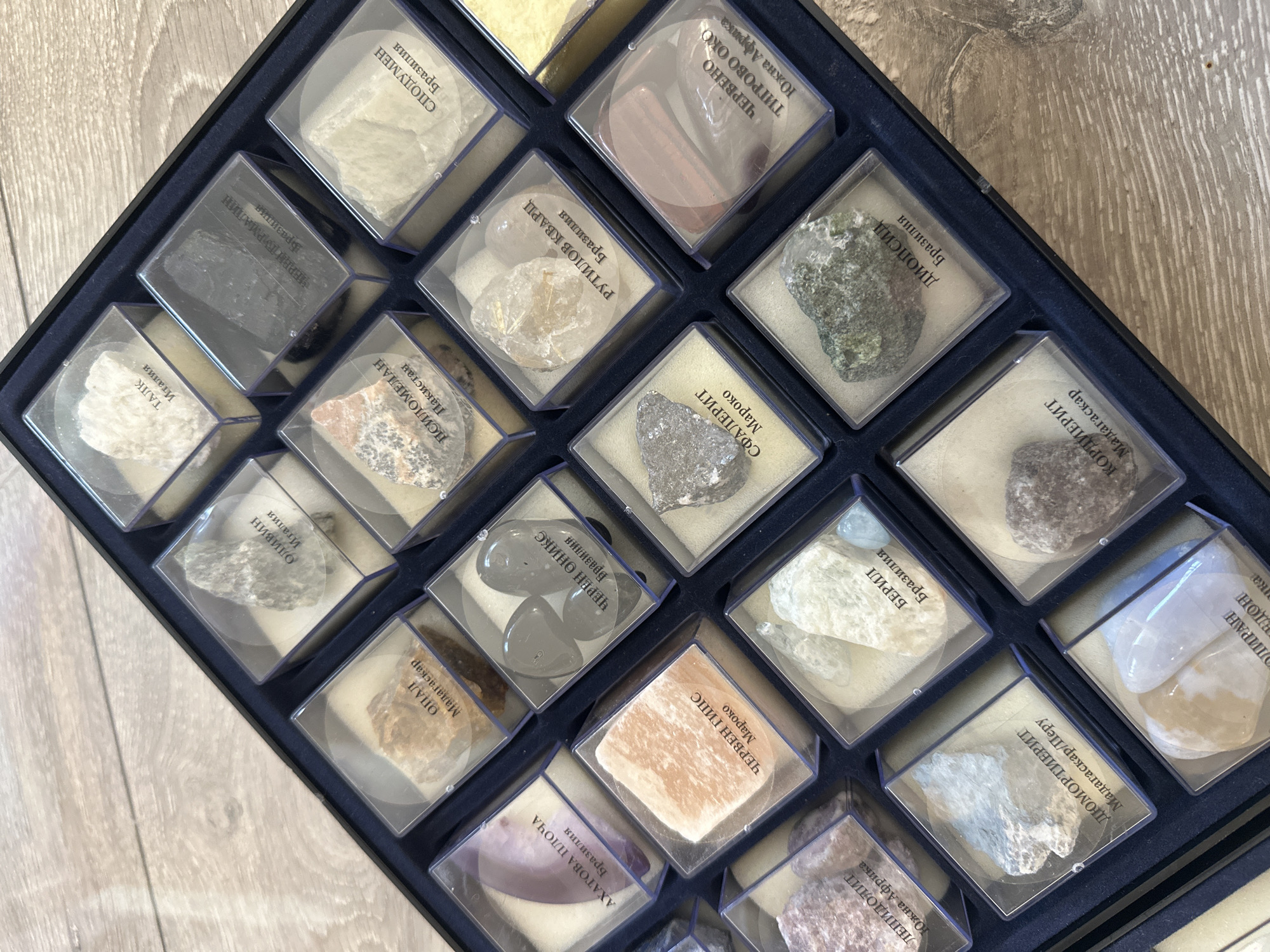 The image shows a collection of mineral samples displayed in a dark blue box. Each sample is contained within a small, clear plastic case. The samples vary in color and texture, ranging from white and translucent to dark gray and opaque. Labels in Cyrillic text are affixed to each case, identifying the mineral and its origin. The box contains minerals such as "LEPIDOLITE", "AGATE SLAB", "OPAL", "OLIVINE", "TALC", "RED GYPSUM", "BLACK ONYX", "PSILOMELANE", "GREEN TOURMALINE", "BERYL", "SPHALERITE", "RUTILE QUARTZ", "SPODUMENE", "OLIGOCLASE", "CORDIERITE", "DIOPSIDE", and "RED TIGER'S EYE". The origins of these minerals are identified as being from various locations, including South Africa, Madagascar, Peru, Brazil, Morocco, Pakistan and Italy. The background is a wooden surface.