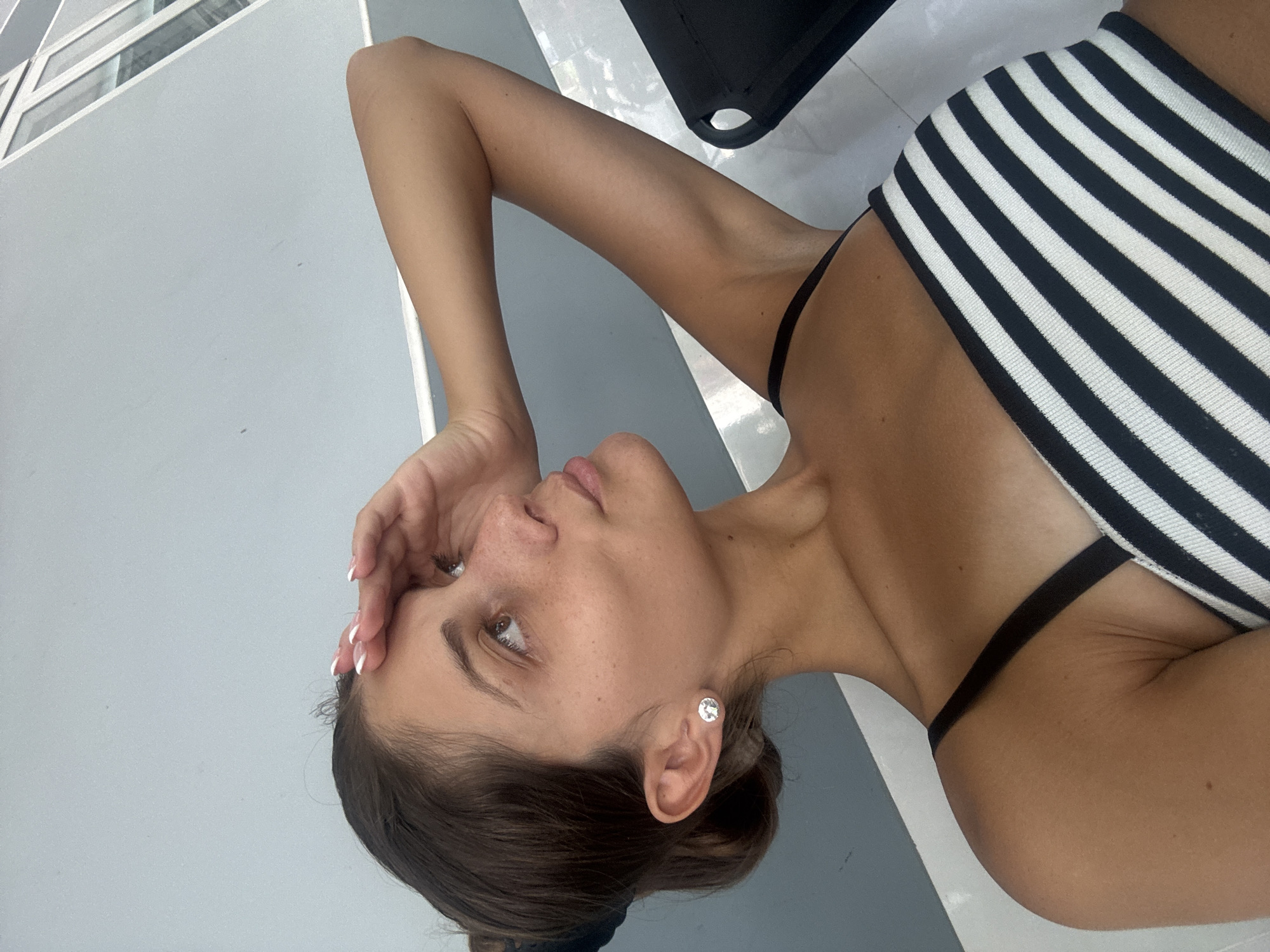 An eye-level shot captures a woman in a reclining or lying down position, looking upwards with a calm, slightly pensive expression. Her body is angled diagonally across the frame, with her head in the bottom right corner and her feet (not visible) towards the top left. She has dark hair, pulled back, and light brown eyes, with visible freckles on her nose and cheeks. Her right hand, adorned with a white-tipped French manicure, is gently resting on her forehead. She wears a silver or clear stud earring in her visible right ear.
The woman is wearing a black and white striped top, likely a bikini top, with a thin black strap visible over her shoulder. Her skin has a sun-kissed tone.
The background consists of clean architectural lines. To her left, a light gray wall or surface dominates, with reflections from a white-framed window structure suggesting an indoor or semi-outdoor setting. To her right, a partial view of a black chair or piece of furniture is visible. The bright, even lighting indicates it is daytime with clear weather. The overall setting appears modern and minimalist, consistent with a contemporary building or resort in a location like Đà Nẵng, Vietnam. No text is visible.