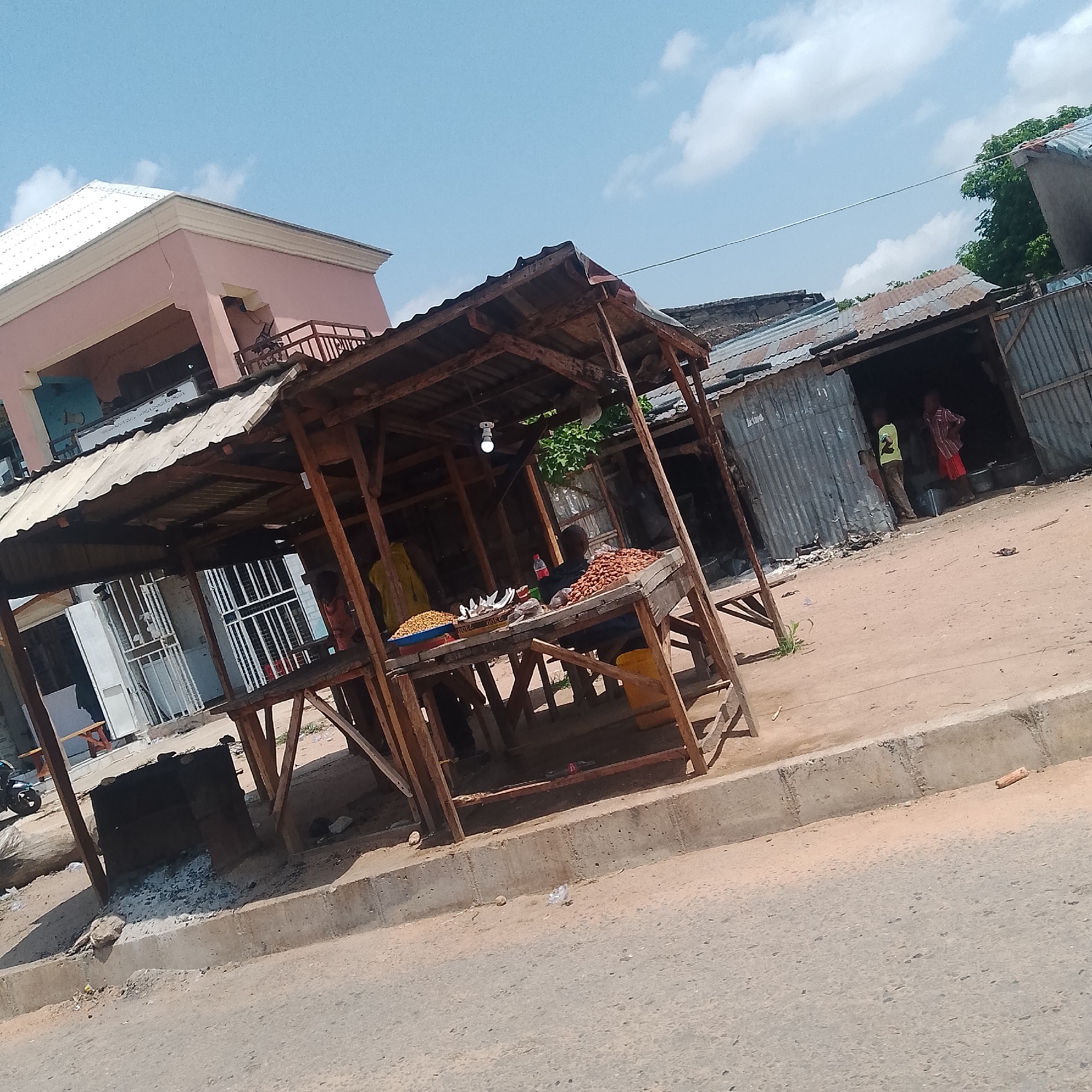 The image depicts an outdoor market scene in Jalingo, Nigeria, under a bright, partly cloudy sky, suggesting daytime. The primary focus is a makeshift wooden stall with a corrugated metal roof, displaying various food items for sale. A person is seated behind the stall, attending to the goods, which include what appear to be nuts or grains in containers, and some bagged items.
Adjacent to this stall, another structure with a similar corrugated metal roof is visible, housing more market activity. Two individuals are seen inside this second structure, one wearing a yellow shirt and the other a plaid shirt and red shorts, suggesting they are vendors or customers. The overall setting is a dusty, open area with a paved road in the foreground and a concrete curb separating the road from the market stalls.
In the background, a light pink building with a white roof stands out, indicating a more permanent structure in the vicinity. The market appears to be functional, with goods laid out and people present, implying a typical day's trade. There is no visible text in the image. The scene conveys a sense of everyday commerce in a Nigerian town.