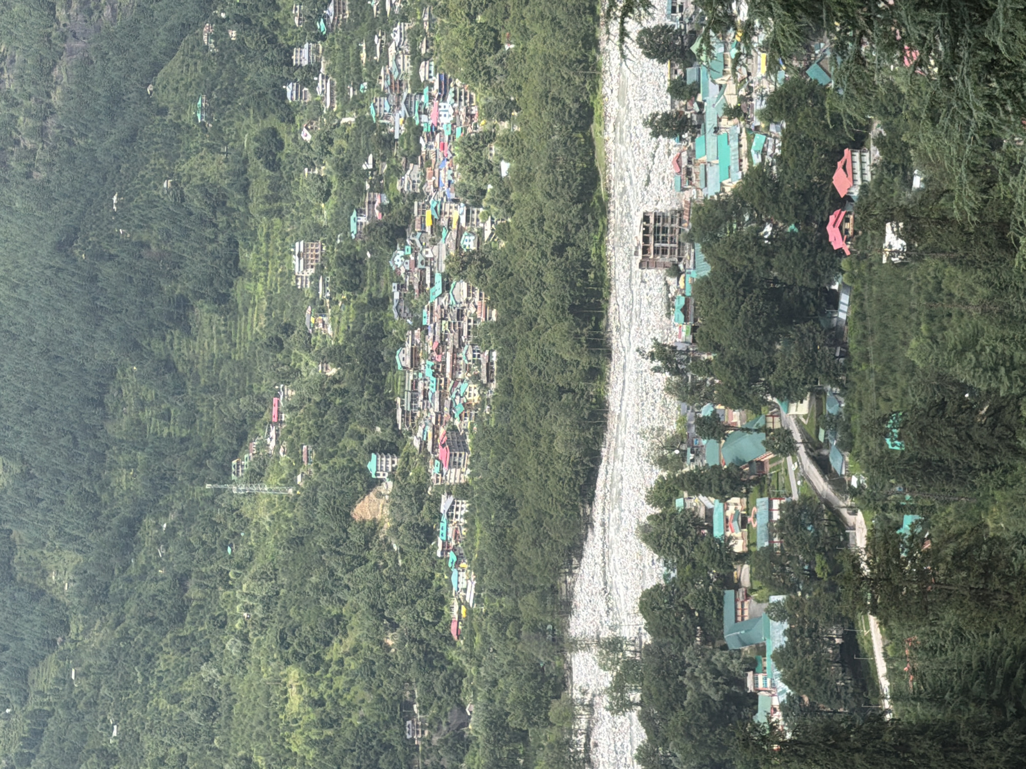 The image depicts a panoramic view of the town of Bahang in India, nestled within a lush green mountainous landscape. The town is characterized by its densely packed houses, many with brightly colored roofs in shades of red, blue, and green, clustered together on the slopes of the hills. A wide, rocky riverbed runs through the valley, bordered by a dense forest of dark green trees. In the foreground, more houses and trees are visible, with a bridge crossing a smaller waterway or road. The overall impression is of a vibrant community integrated into a natural, hilly environment.
