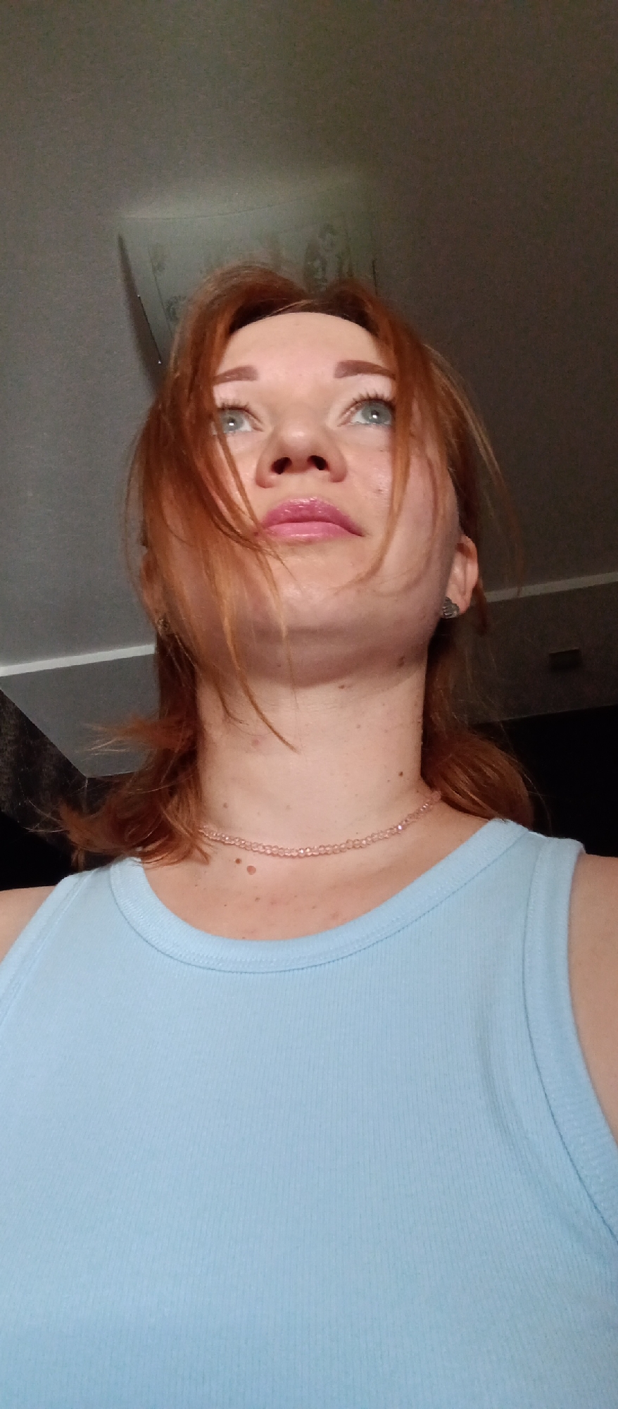 A woman with auburn hair and a light blue tank top is looking upwards. She is wearing a pink beaded necklace. The background is out of focus, but appears to be an indoor setting with a light fixture on the ceiling. The location is Unknown, Unknown.
