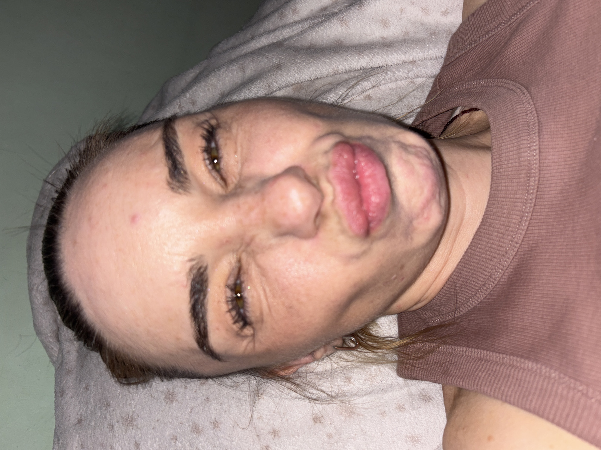 A close-up, waist-up, indoor, slightly tilted, selfie of a fair-skinned Caucasian woman with auburn hair pulled back. She is lying down, resting on a white, patterned blanket with a grey blanket draped behind her head and shoulders. The woman is making a puckering face with her lips protruding and her eyes squinting. She is wearing a pink ribbed tank top. She has dark eyebrows, visible freckles, and is wearing a silver hoop earring in her left ear. The background consists of a pale green wall. The lighting is dim and appears to be artificial.