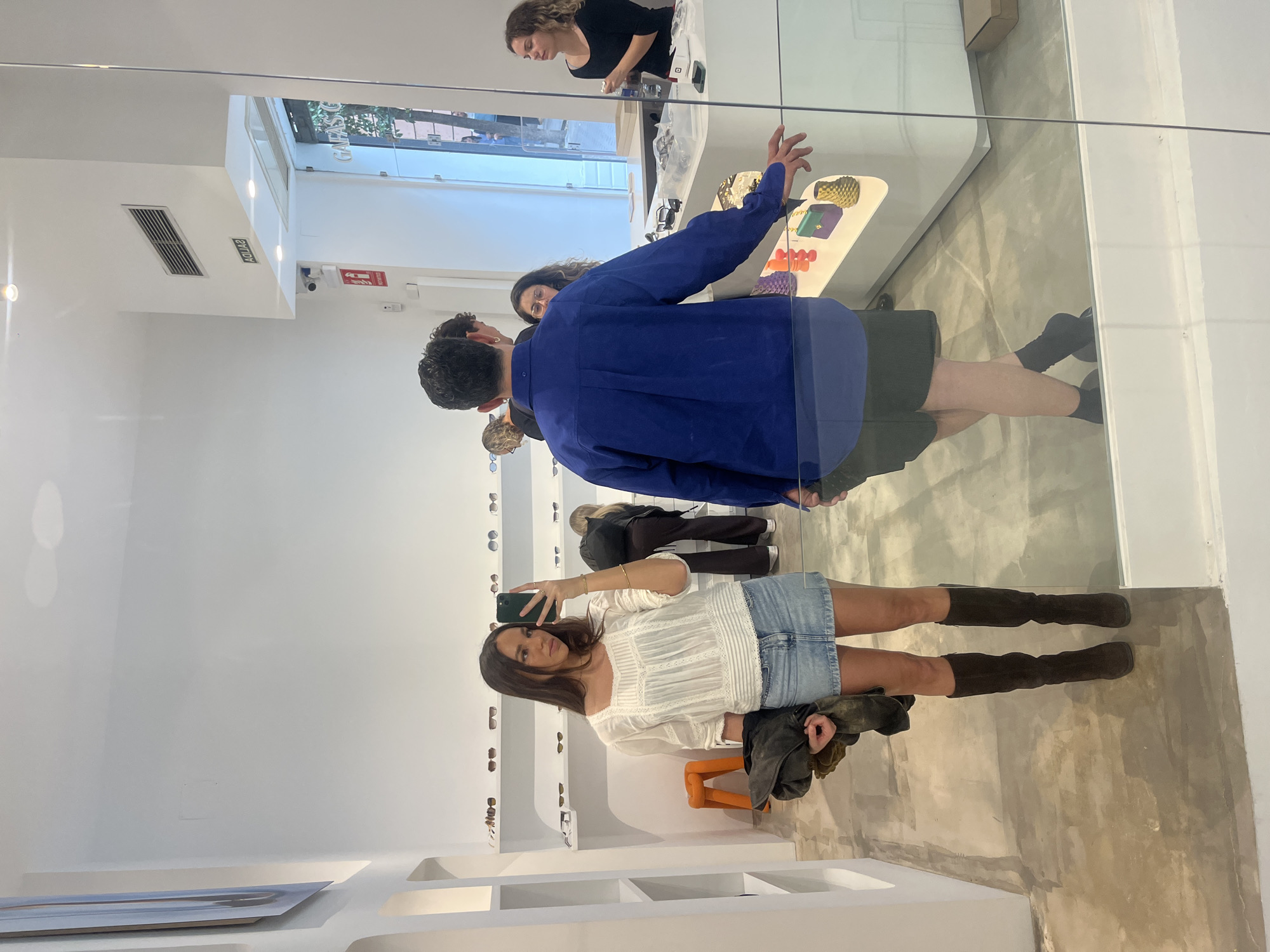 A woman takes a selfie in a shop, the reflection is of customers and staff. The environment appears to be a modern retail space, likely in Madrid, Spain. A woman in a white top and denim skirt takes a selfie with her phone. She is wearing tall brown boots. A jacket sits on an orange stool next to her.
Behind her, mirrored in the glass, is the interior of the shop. There are shelves displaying glasses and eyewear on the left-hand side. In the background, there is a male customer in a blue shirt browsing with a group, and a female staff member working at the counter. A selection of objects rests on the counter.