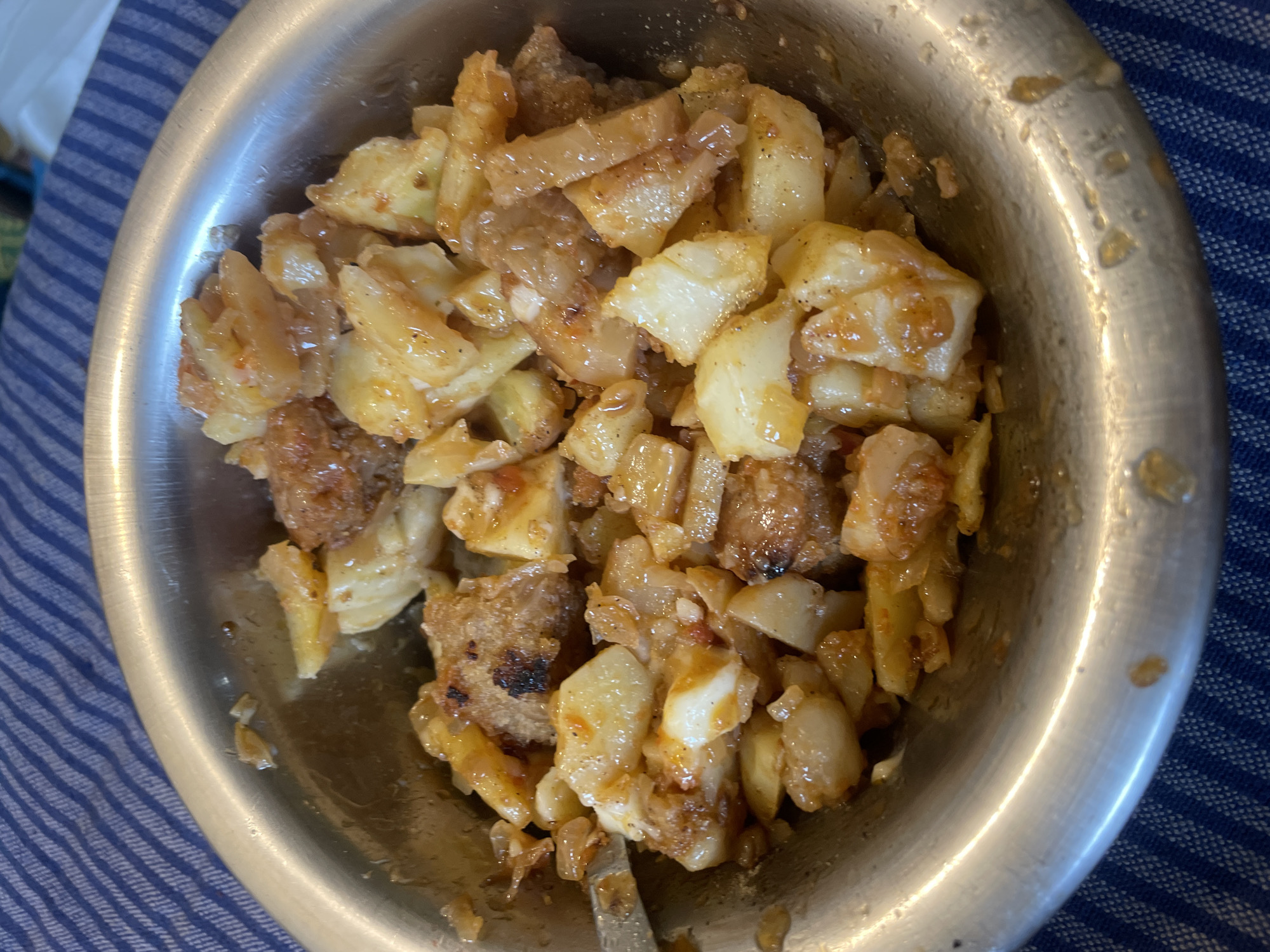 A close-up, overhead shot shows a metallic bowl filled with a dish of diced potatoes and meat. The potatoes are cut into bite-sized cubes and appear to be cooked in a sauce that gives them a glossy, slightly orange hue. Interspersed with the potatoes are pieces of meat, some appearing to be ground or finely chopped, also coated in the sauce. The dish is resting on a blue and white striped fabric. The lighting suggests an indoor setting, likely a meal being served. The specific location within Haini, India, cannot be determined from the visual information provided.