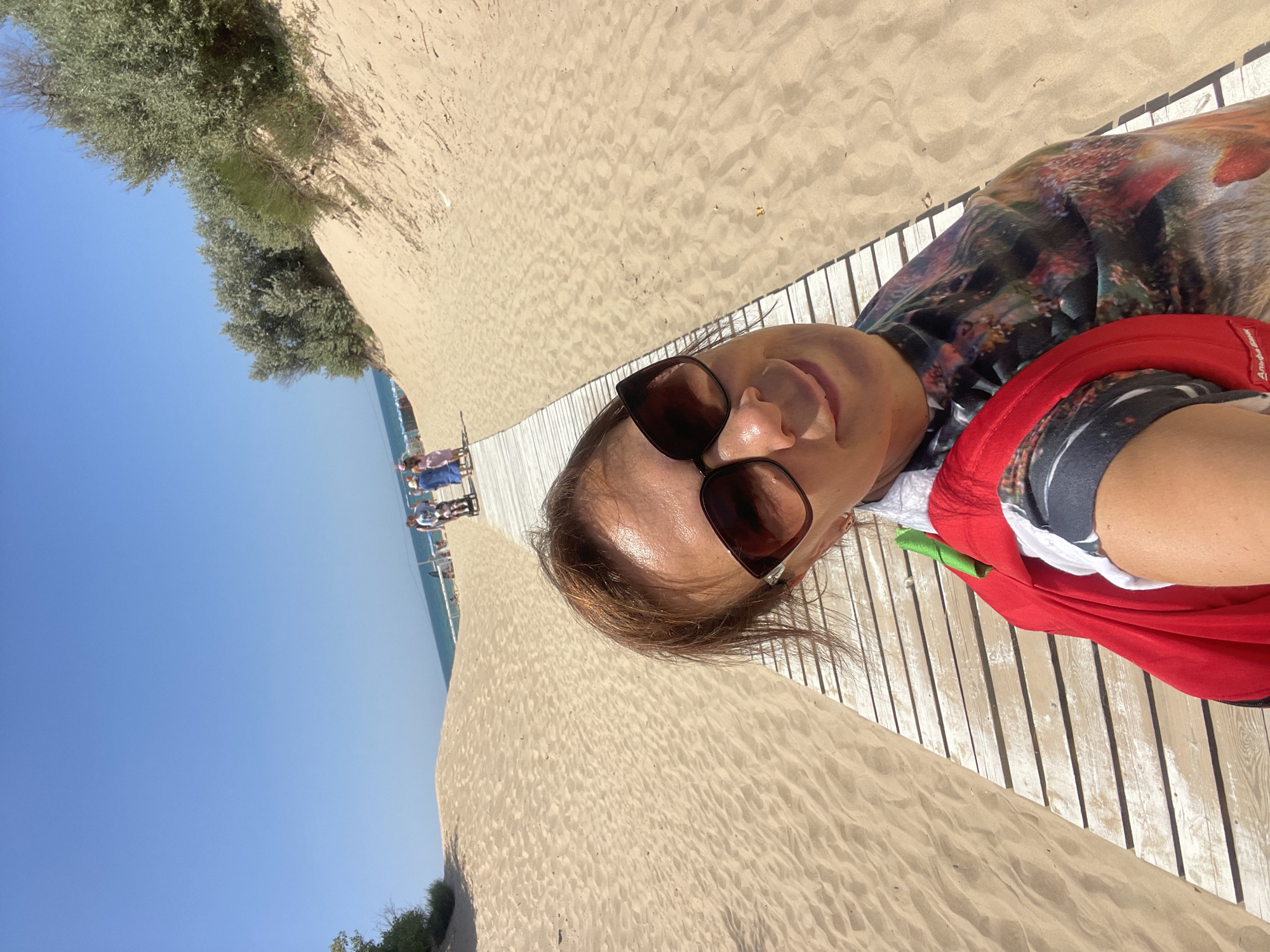 A woman with sunglasses and a red backpack is taking a selfie on a wooden boardwalk at a sandy beach. In the background, a group of people are walking along the boardwalk toward the sea, and beach umbrellas are visible. The sky is clear and blue. The scene is likely located in Anapa, Russia, a popular resort town on the Black Sea.