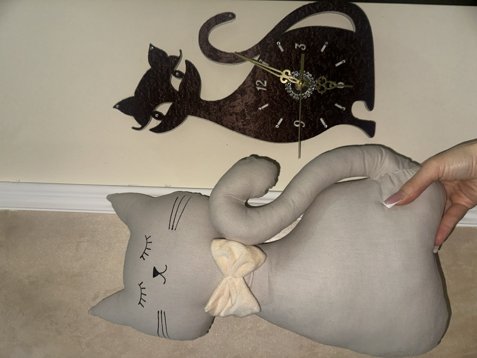 An interior scene in Yerevan, Armenia, displays a large, light-grey fabric cat plushie positioned to the left of a dark brown cat-shaped wall clock, both against a light beige wall. A human hand, with manicured nails featuring light-colored tips, supports the base of the plushie from below. The plushie has embroidered closed eyes with eyelashes, a black nose, whiskers, and a light peach-colored bow tied around its neck, with an elongated body and a curved tail. The wall clock, crafted from dark brown material with a textured appearance, is a stylized cat silhouette. It features white numerical markers (9 through 3 visible) and smaller minute markers, with gold-colored hour and minute hands emanating from a central, jewel-studded decorative element. A vertical white trim piece is visible on the far left of the frame.