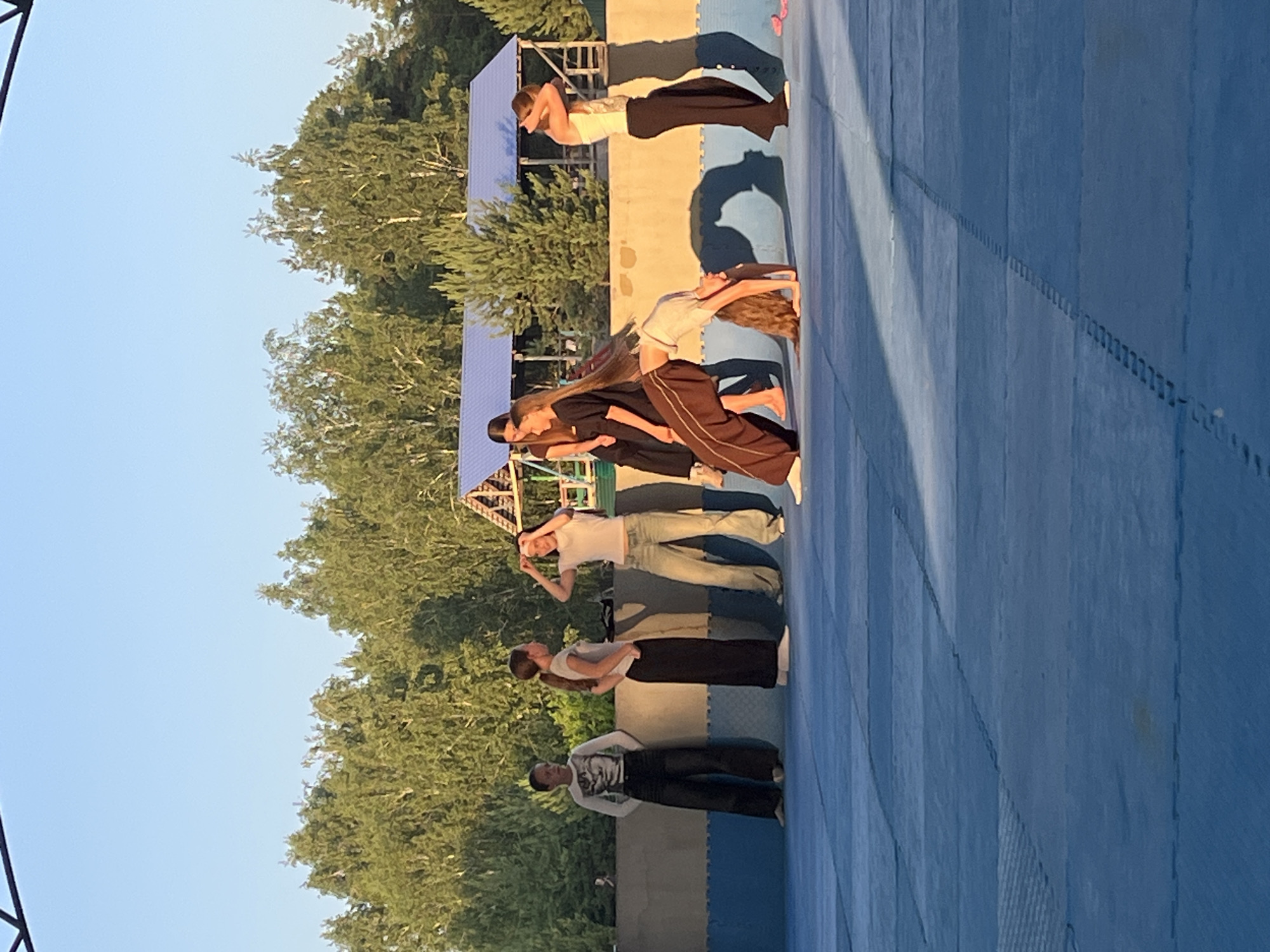 This image captures a group of people, likely teenagers, engaged in a physical activity on a blue matting surface. The subjects are positioned in a line, performing what appears to be a synchronized dance or gymnastics routine, characterized by arched backs and bending poses. The scene is outdoors, with a clear blue sky overhead and lush green trees visible in the background. The lighting suggests it is daytime, likely late afternoon or early evening, given the long shadows cast by the individuals.
The individuals are dressed in casual athletic wear. One person wears a white t-shirt and dark pants, another a white t-shirt and jeans, and a third person is in a black top and brown pants. Further down the line, other participants are visible in similar attire. The blue matting has a grid-like pattern, indicating it's a designated area for activities like sports or performances. A structure, possibly a small stage or seating area, is partially visible behind the trees. The overall atmosphere appears to be focused and collaborative, with the participants concentrating on their movements. The location context provided is Krasnoturansk, Russia. There is no visible text in the image.