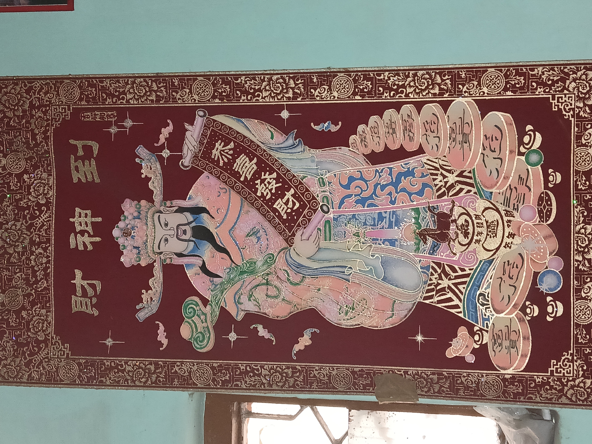 The image is a close-up of a rectangular, maroon and gold-colored artwork depicting a deity or mythical figure, potentially of East Asian origin. The figure is illustrated in profile, holding a scroll-like object with Chinese characters. The artwork features intricate patterns and designs, including stylized coins and floral motifs along the edges. It is mounted on a light turquoise-colored wall, with a portion of a window visible at the bottom. The window frame appears to be made of wood. The location is Bengaluru, India.