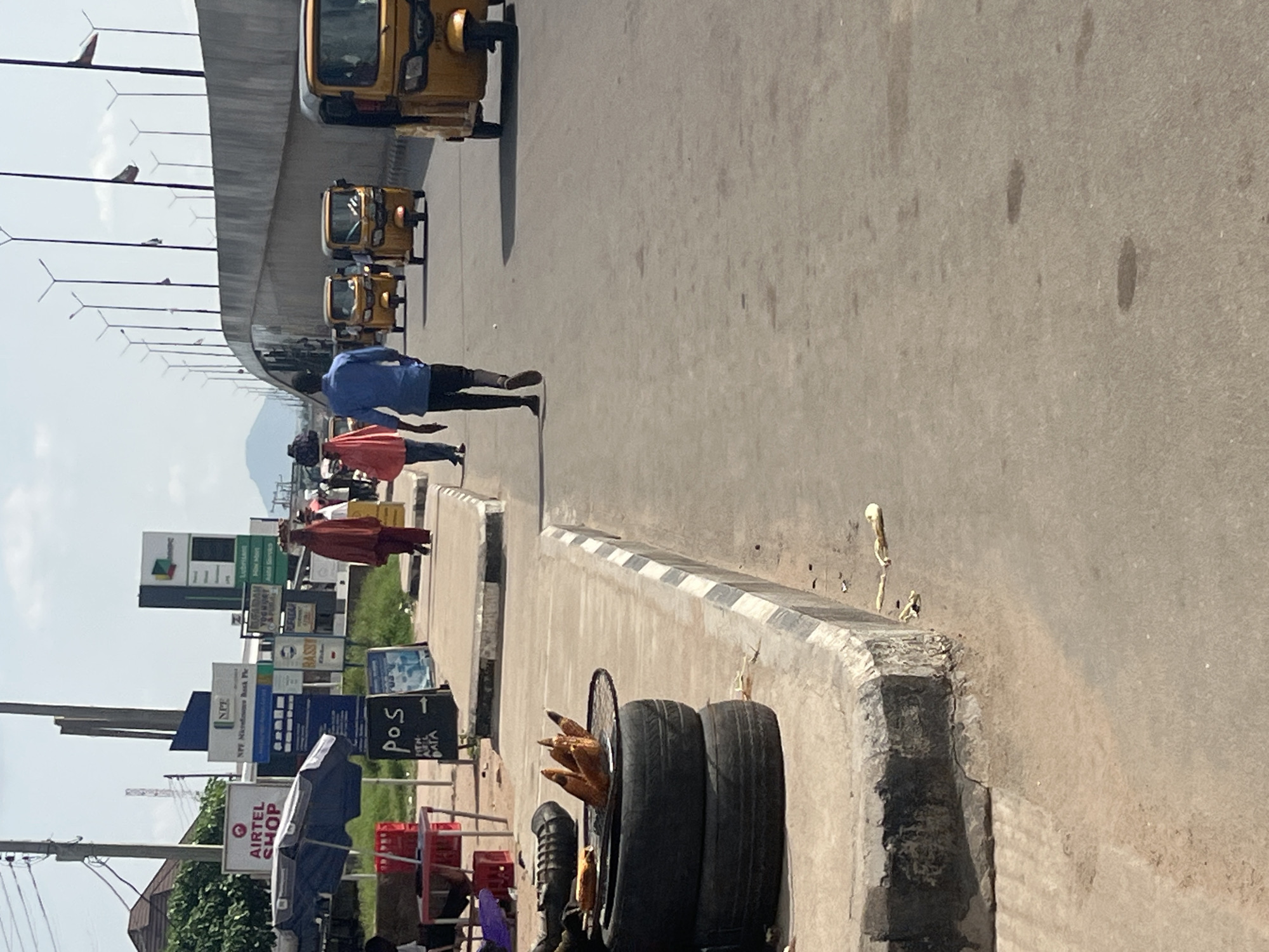 The image depicts a street scene in Jalingo, Nigeria, likely near a commercial area. In the foreground, a makeshift grill for roasting corn, made from stacked tires, is visible on the sidewalk. Adjacent to it, a POS (Point of Sale) sign is displayed. The sidewalk is lined with a curb.
Further down the street, several people are walking along the sidewalk. Two individuals are particularly noticeable: one in a red or maroon garment and another in a blue shirt and black trousers. They are walking away from the viewer. Several yellow "keke napep" (three-wheeled motorized rickshaws) are on the road, some moving and one parked.
In the background, various signs are mounted on poles, including one for "AIRTEL SHOP" and another indicating "NPP Microfinance Bank Plc." There are also utility poles with electrical wires. The environment appears to be an urban setting with a road, sidewalks, buildings, and overhead infrastructure. The sky is visible, with some clouds. The overall impression is one of a bustling commercial street in a Nigerian city.