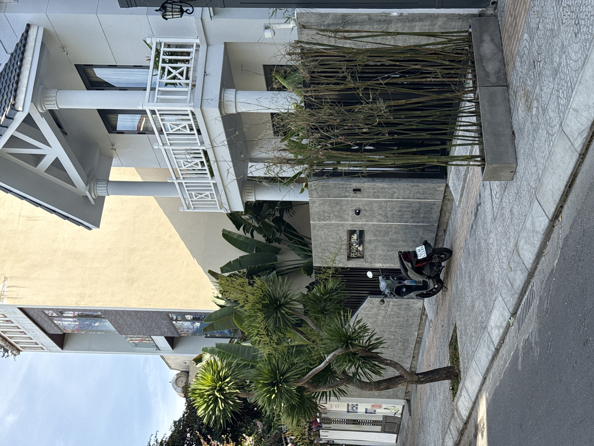 This is a street-level, medium shot taken in Da Nang, Vietnam. The scene depicts a residential area with a modern, multi-story house and a neighboring building. The house on the right is white with a white balustrade on the second-floor balcony supported by columns. A dark-colored motorcycle is parked on the sidewalk in front of a grey concrete wall. A small tree with spiky green leaves stands to the left of the motorcycle, and a planter box filled with tall bamboo stalks is situated to the right of the wall. The pavement is made of grey concrete tiles. The sky is clear and blue.