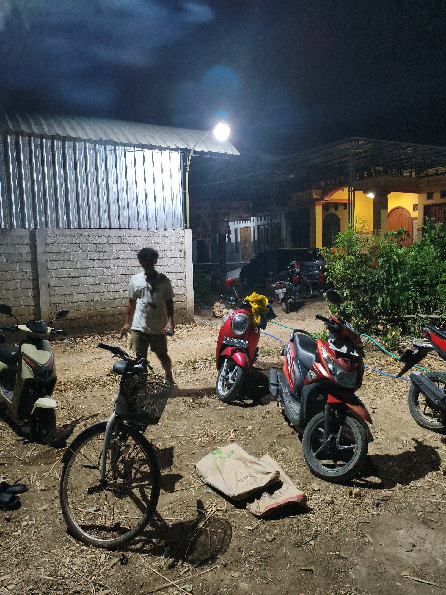 The image depicts an outdoor scene at night in the city of Karangtalun, Indonesia. In the foreground, a person wearing a light-colored t-shirt and shorts stands amidst several parked motorcycles and a bicycle. The ground is covered with dirt, dry leaves, and debris. To the left, a cream-colored scooter is parked next to a white brick wall. A bicycle with a basket is positioned in the center of the frame. In the middle ground, two red scooters are parked, one of which has a yellow cloth draped over its handlebars. Behind the scooters, a dark car is visible, and further back, a building with yellow walls and a dark roof is illuminated by interior lights. To the left, a long, corrugated metal structure with a slanted roof is present, and smoke or mist is rising above it. The overall impression is a residential or utilitarian outdoor area at night, with multiple vehicles suggesting it may be a place of residence or a temporary parking area.