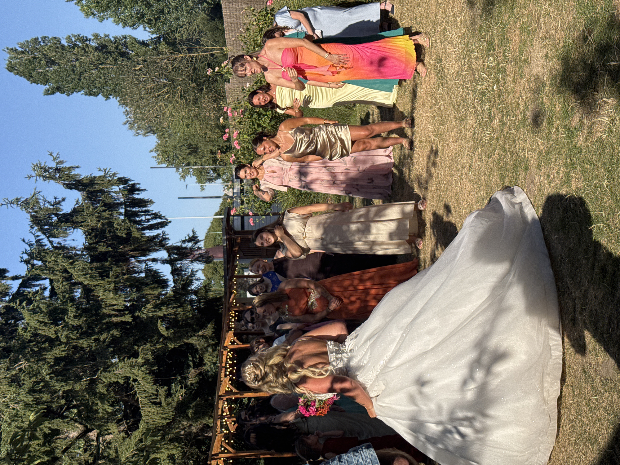 The image depicts a wedding party gathered outdoors in what appears to be a garden setting. The bride, in a voluminous white gown, stands facing away from the camera, her back adorned with intricate white floral details. She holds a bouquet of colorful flowers. Surrounding her are several women, presumably bridesmaids and guests, dressed in various colored gowns and dresses. The attire ranges from a shimmering gold mini-dress and a long, flowing orange dress to pastel pink and yellow floor-length gowns, and a bright, multi-colored pink, orange, and yellow ombre dress.
The environment is characterized by lush green trees providing shade, a clear blue sky, and dry, sun-drenched grass. In the background, a wooden pergola decorated with string lights suggests a venue for celebration or reception. There are also rose bushes visible in the middle ground. The lighting indicates it is daytime, likely late afternoon due to the long shadows.
While the image is rich in visual detail, there is no specific information within the image itself to pinpoint the location to Hem, France. The scene is a generic outdoor wedding celebration.